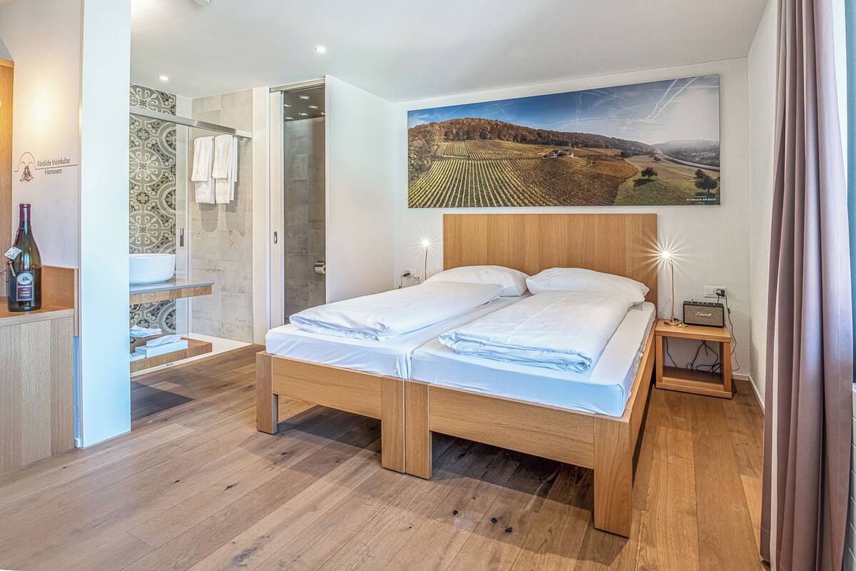 A modern hotel room with a wooden bed, a shower area, a sink, and a large landscape painting on the wall.