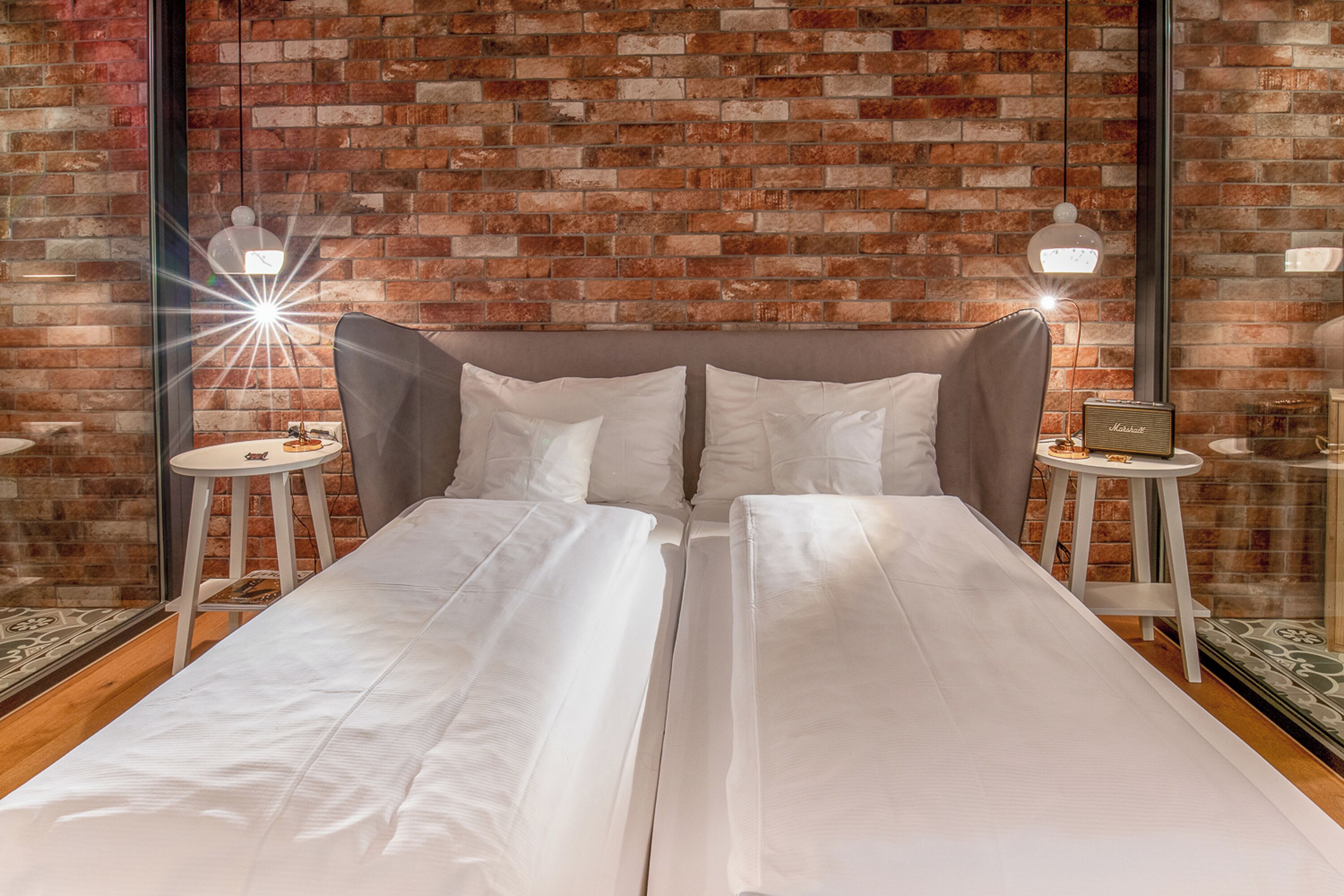 Cama doble con sábanas blancas en una habitación con pared de ladrillo y dos mesitas de noche con lámparas.