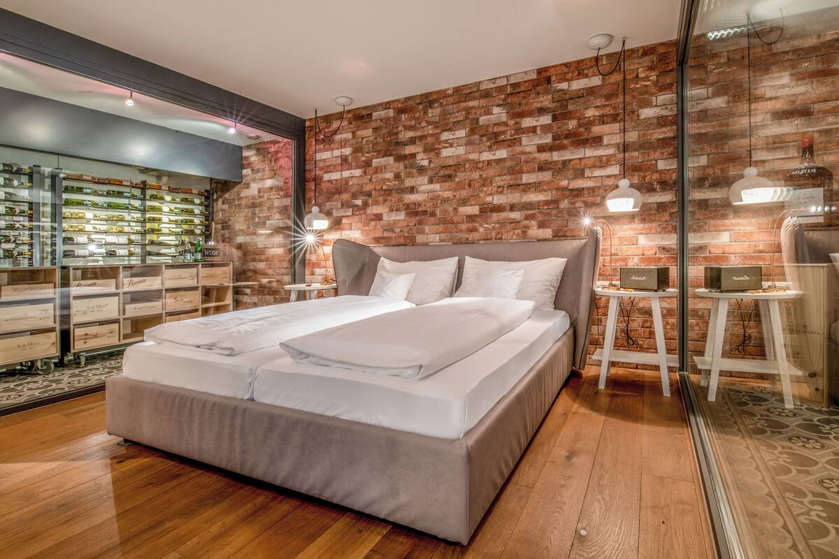 A modern bedroom with a brick wall, a large bed, and a glass display of wine bottles.