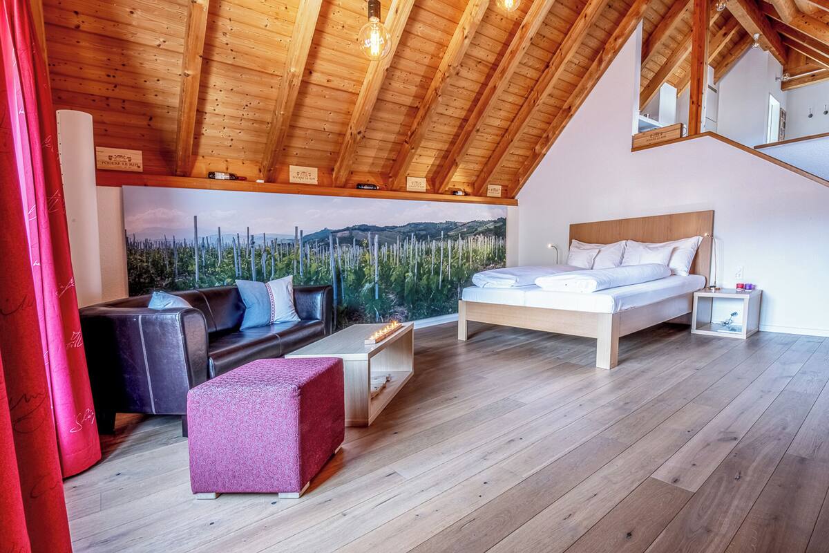 A bedroom with a wooden ceiling, a bed with white bedding, a nightstand, a red ottoman, and a painting of a forest.