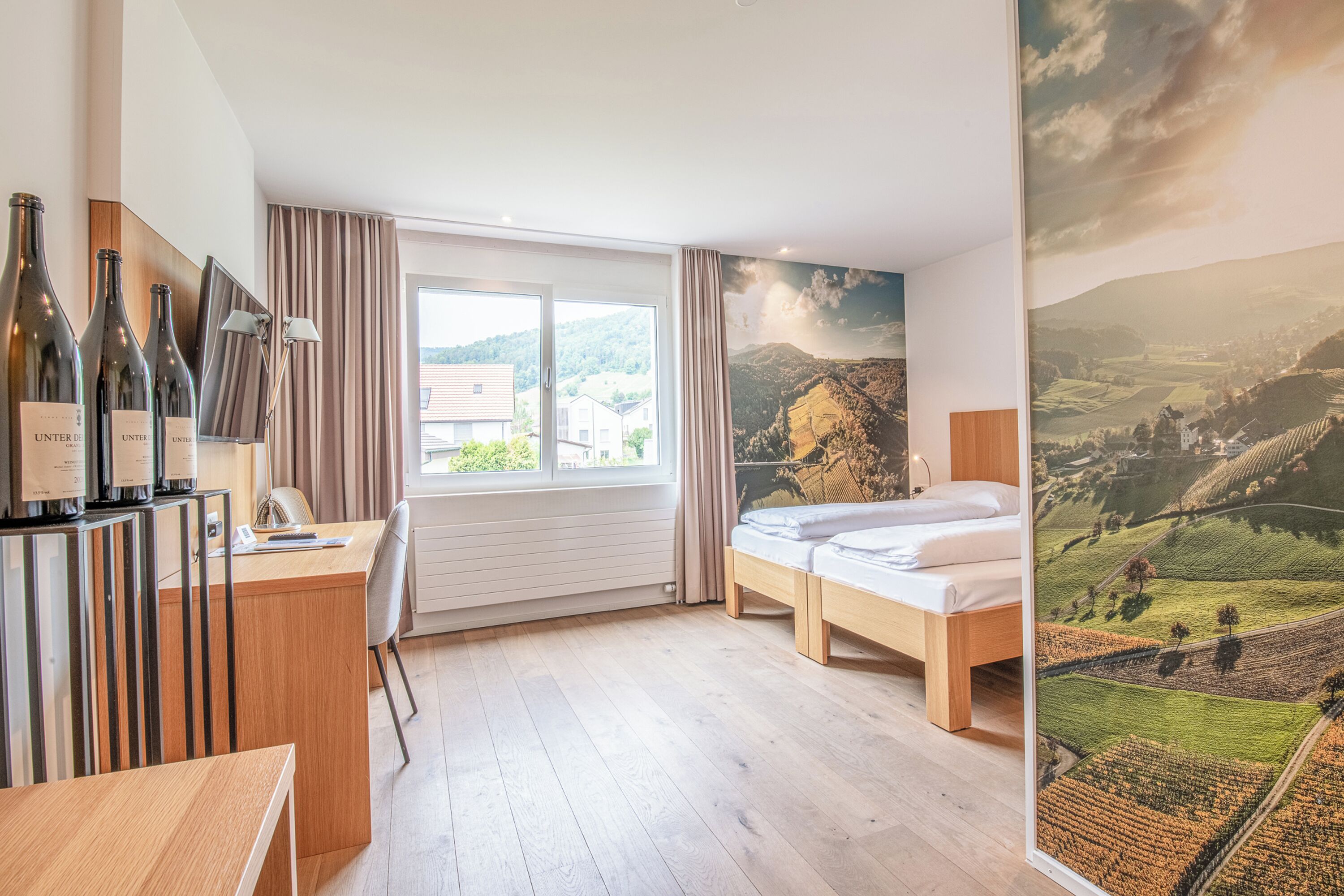 Habitación de hotel con cama, un escritorio con botellas de vino, vistas al campo a través de una ventana y un mural de un paisaje.