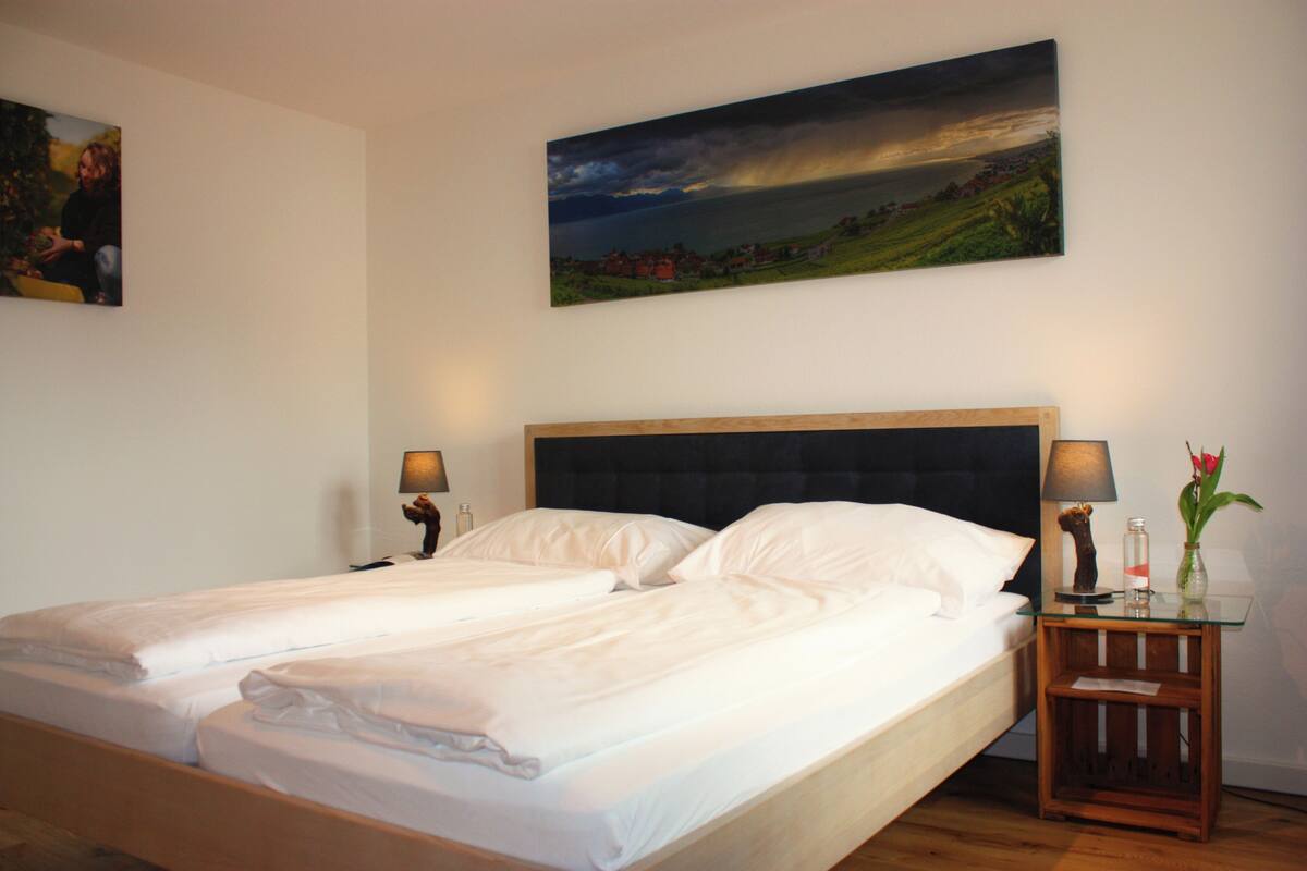 A double bed with a wooden headboard, a bedside table with a lamp, and a painting of a landscape on the wall.