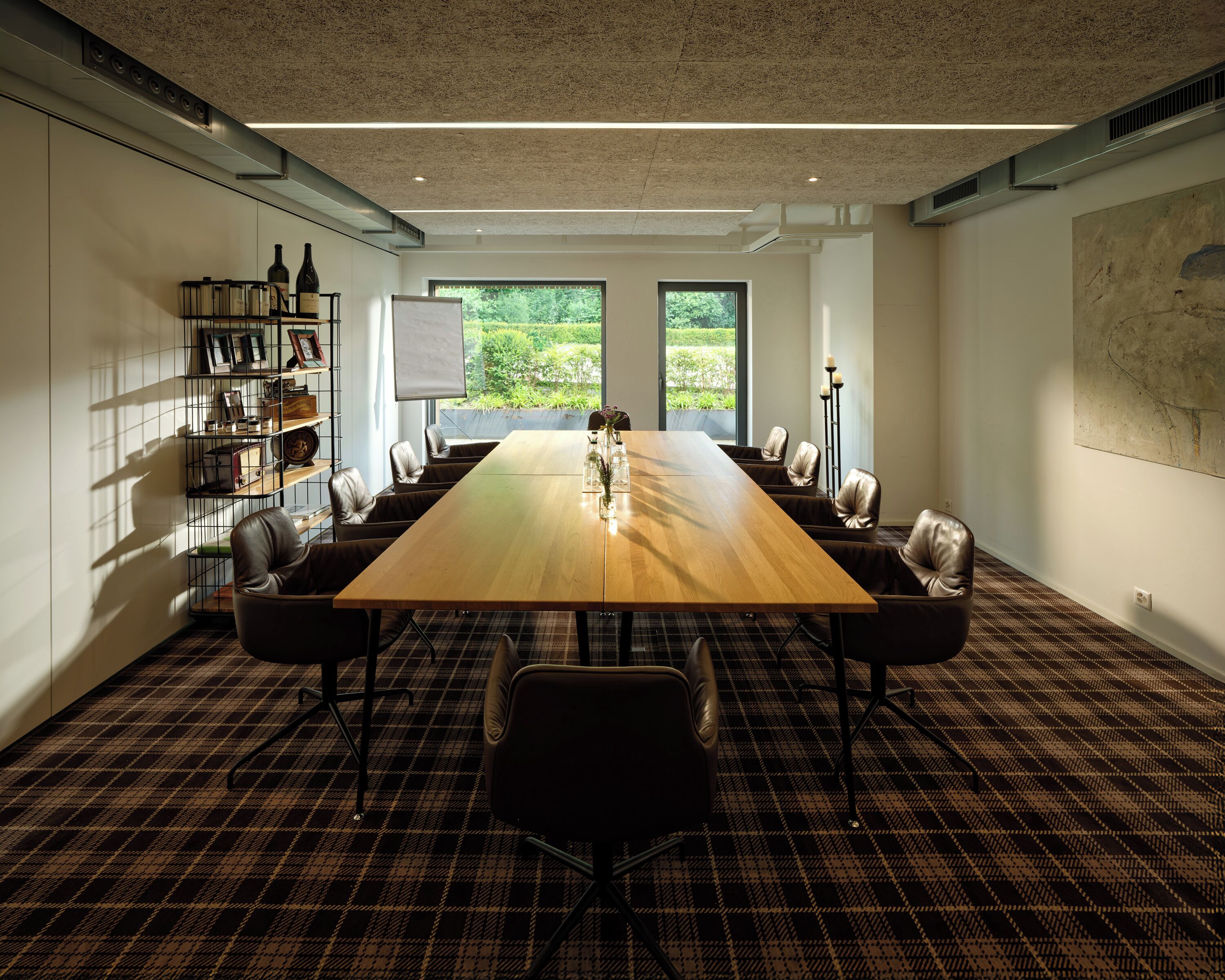 A conference room with a long wooden table, leather chairs, a bookshelf, and a large window with a view of greenery.