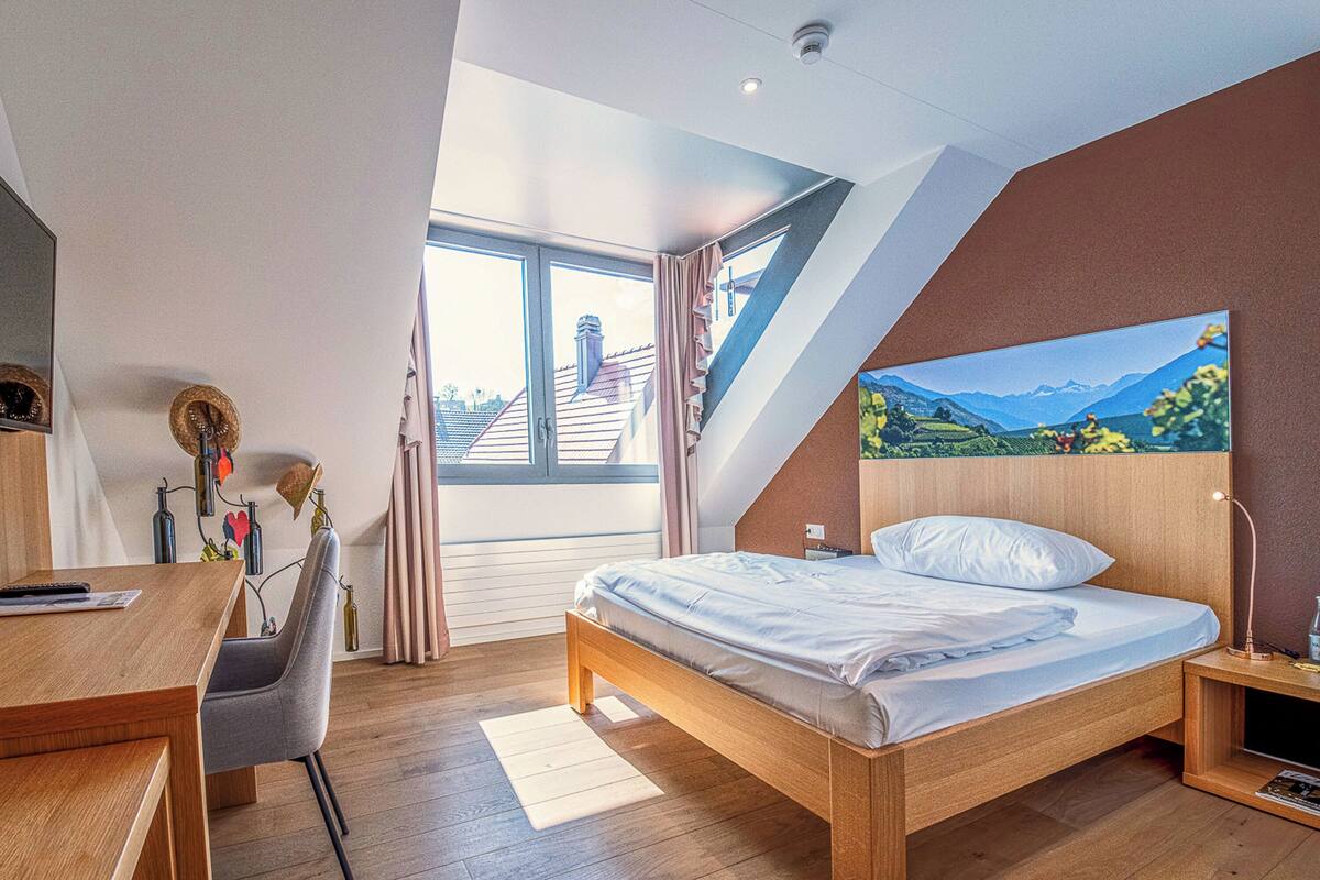A modern bedroom with a wooden bed, a desk, and a large window offering a view of buildings and mountains.