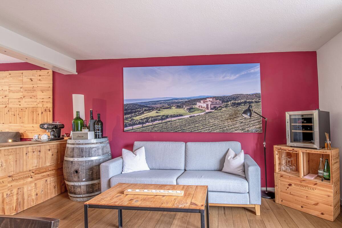 A living room with a red wall, a grey sofa, a wooden coffee table, and a large wine bottle on a barrel.
