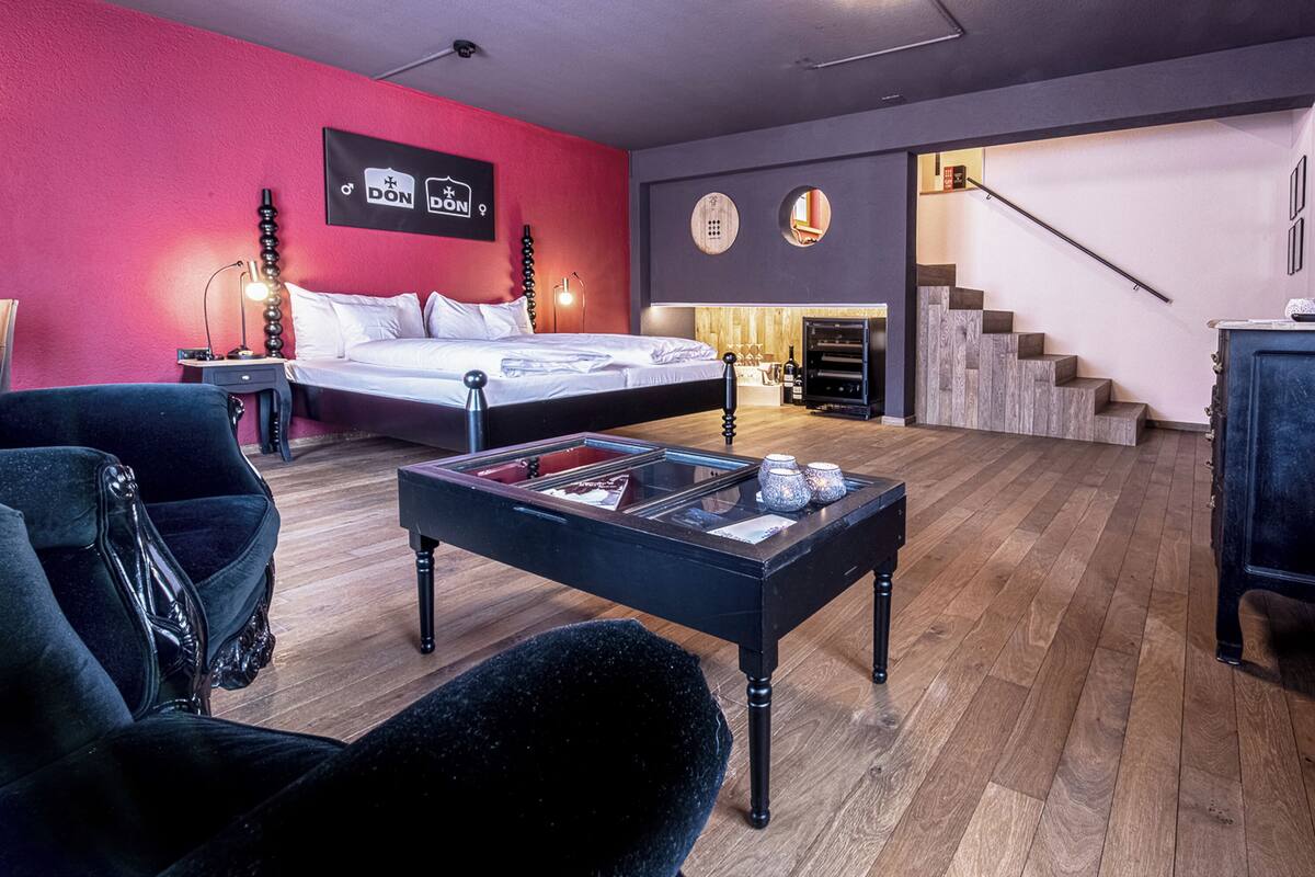 A modern hotel room with a bed, a black coffee table, a red accent wall, and a staircase.
