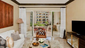 Uma sala de estar espaçosa com um sofá branco, uma mesa de centro com objetos decorativos, uma TV sobre um suporte de madeira e uma varanda com vista para um edifício alto e mobiliário de exterior.