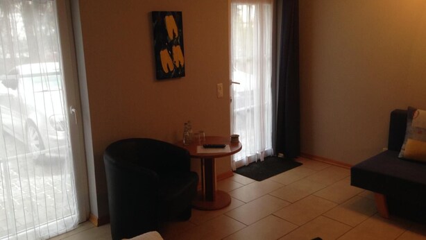 Un salon avec un fauteuil noir, une petite table ronde sur laquelle se trouve une télécommande, et un canapé avec un coussin.