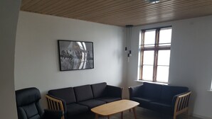 Una sala de estar moderna con un sofá de cuero negro, una mesa de centro de madera y una silla de oficina negra. Un cuadro abstracto de gran tamaño cuelga de la pared, y hay una ventana con persianas a la derecha.