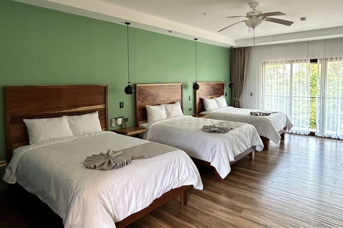 A hotel room with three single beds, wooden headboards, and a large window with curtains.