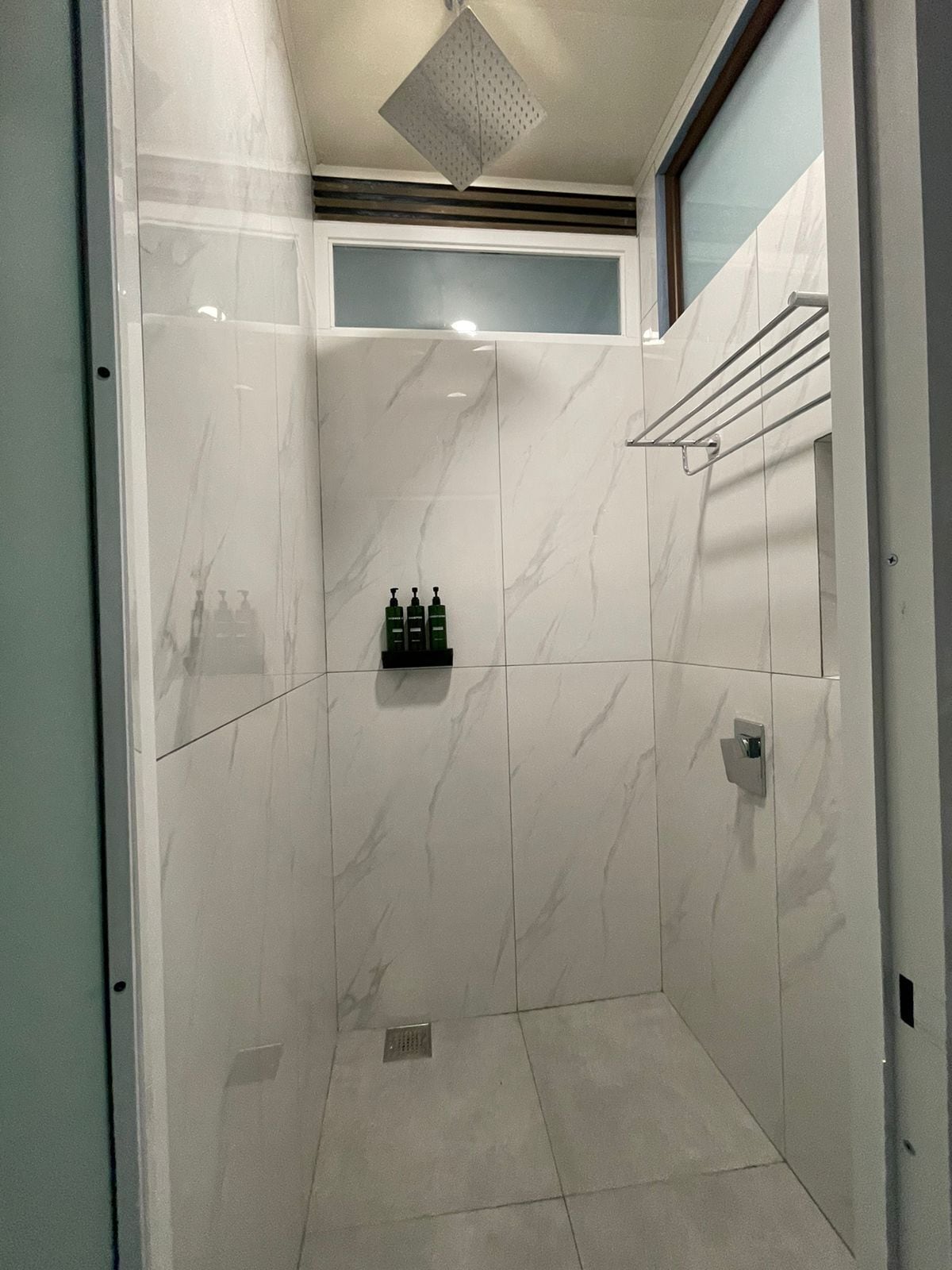 A modern bathroom with white marble tiles, a shower area, and a small shelf with toiletries.