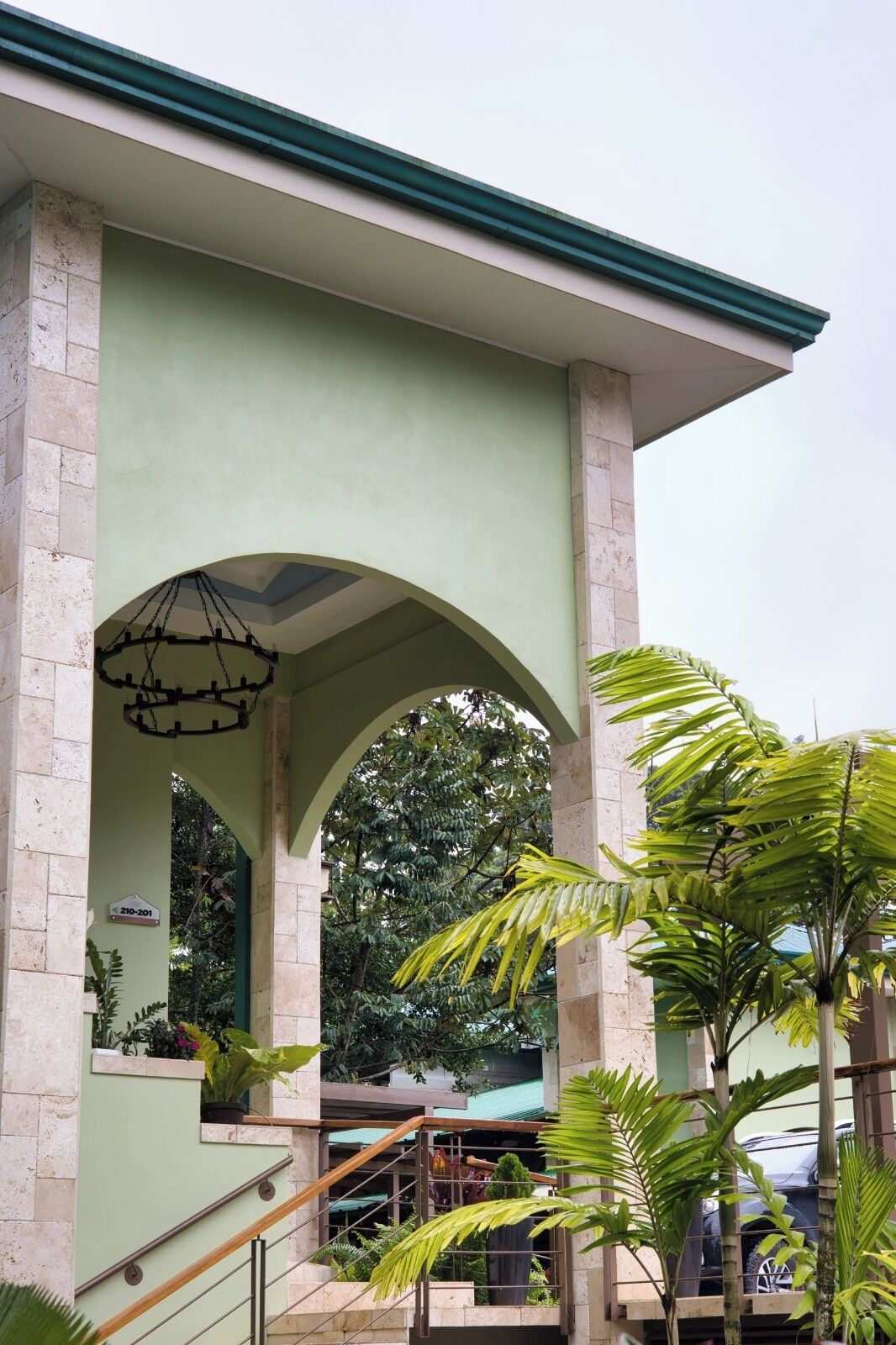 A building with a green facade, a covered entrance with a wooden staircase, and surrounding lush greenery.