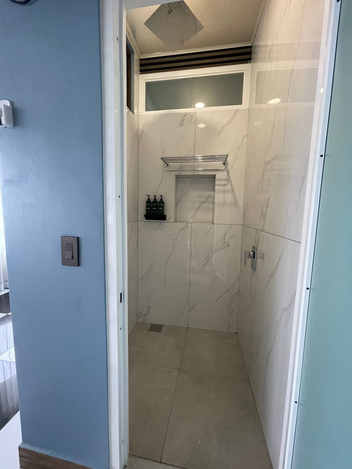 A bathroom with white marble tiles, a shower area, and a small shelf with toiletries.