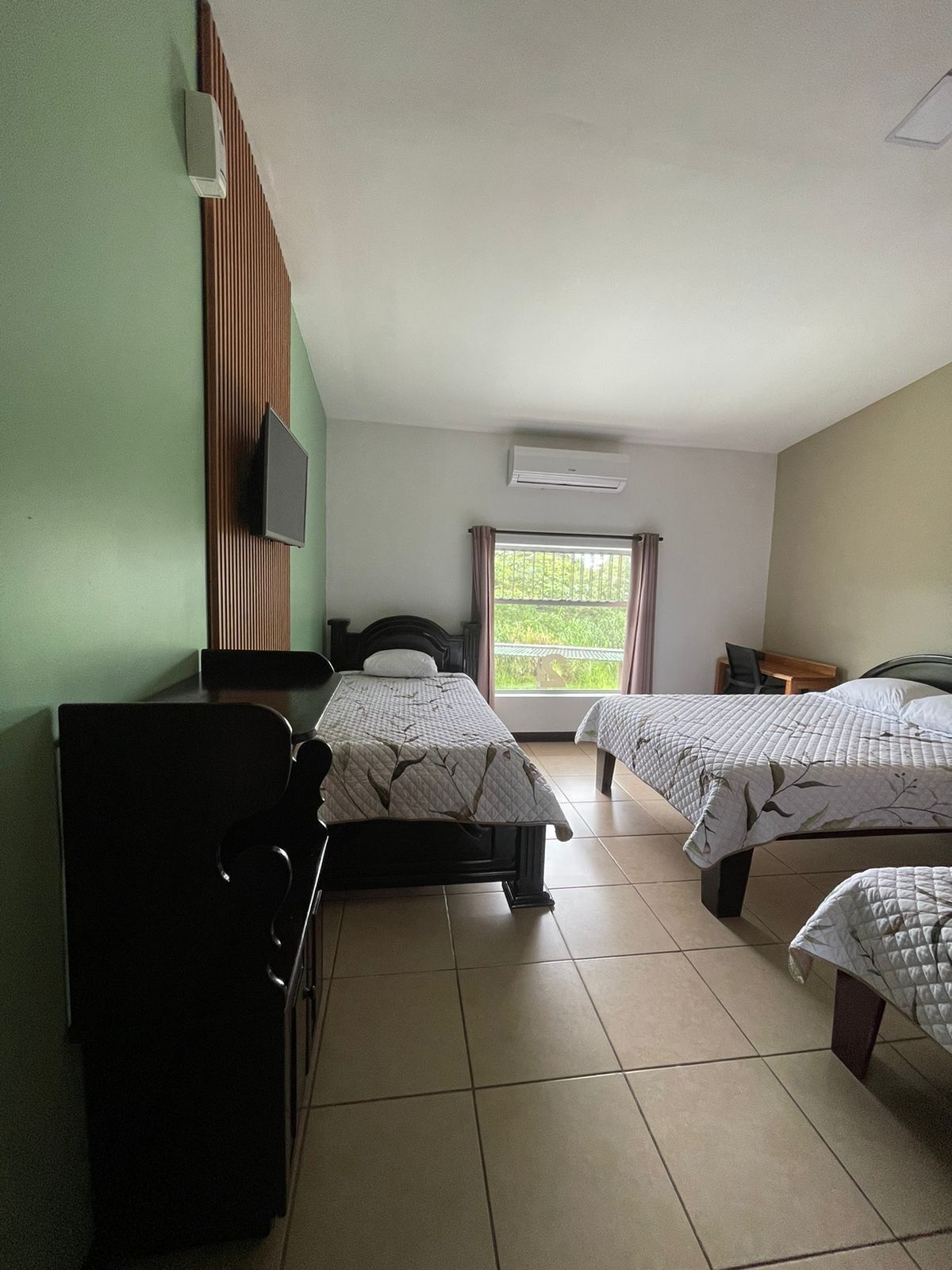 Hotelkamer met twee bedden, een televisie, een airconditioner en een raam met uitzicht op groen.