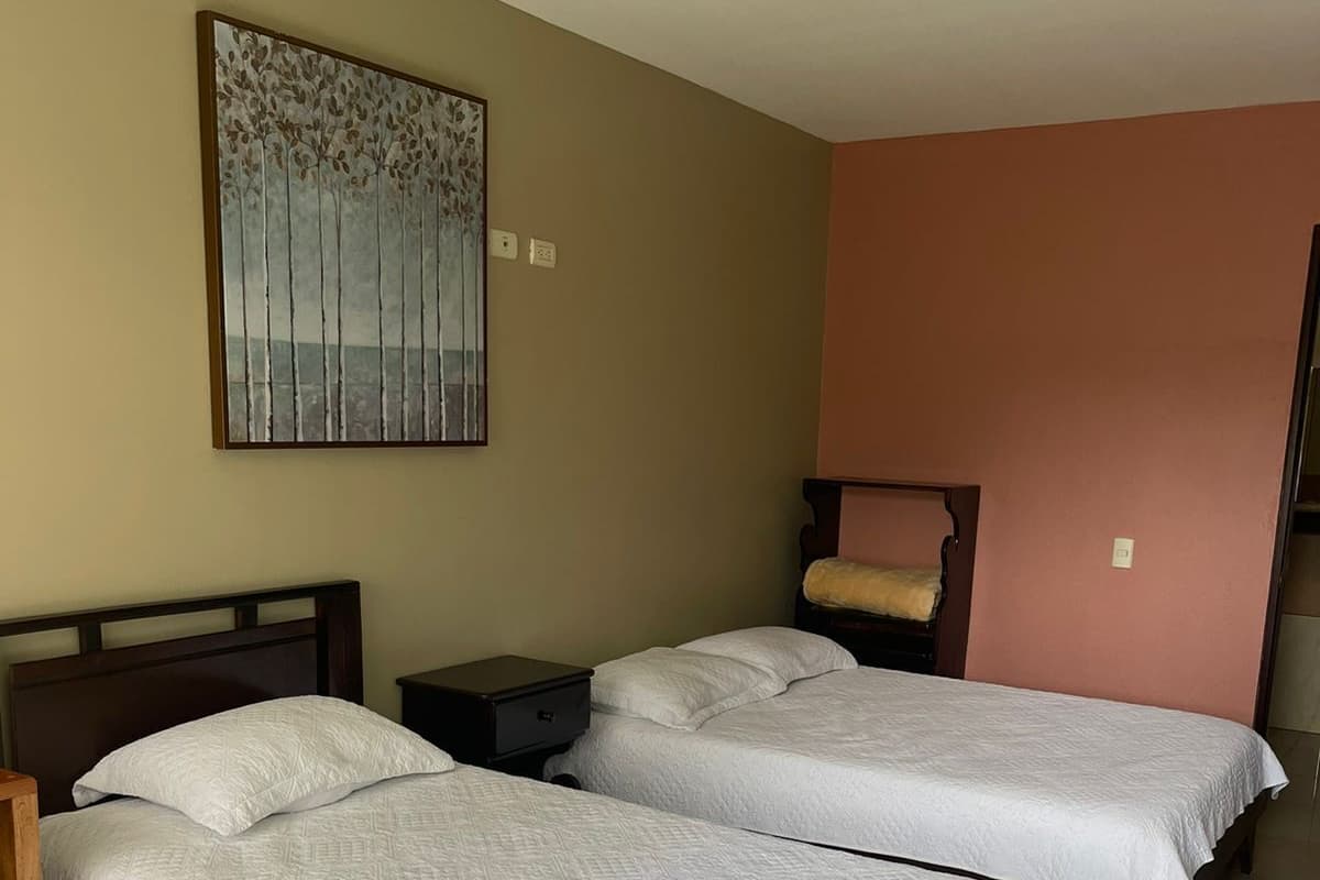 A hotel room with two single beds, a nightstand, a wall-mounted artwork, and a door to another room.