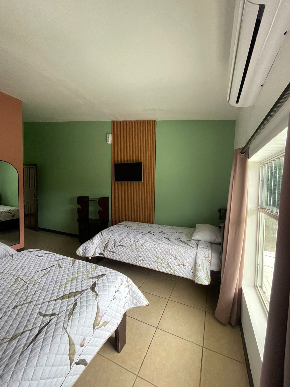 A room with two beds, a television mounted on a wooden panel, and a window with curtains.