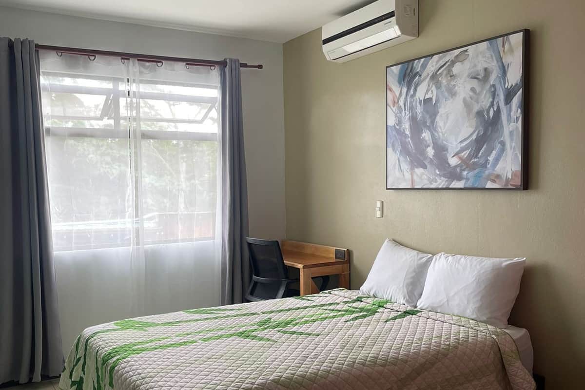 A hotel room with a bed, a desk, and an air conditioning unit.