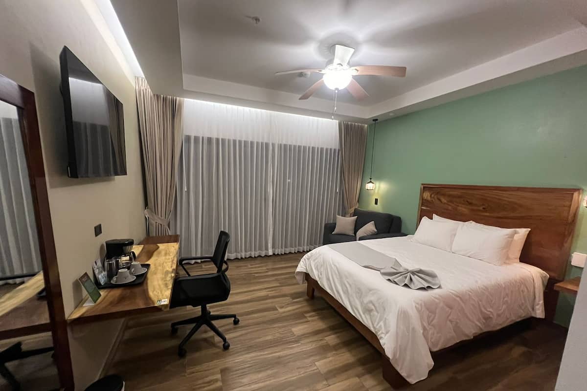 A hotel room with a bed, a desk with a chair, a television, a ceiling fan, and a window with curtains.