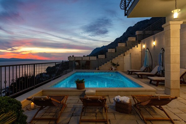 A pool area with lounge chairs, a staircase, and a view of the mountains at sunset.