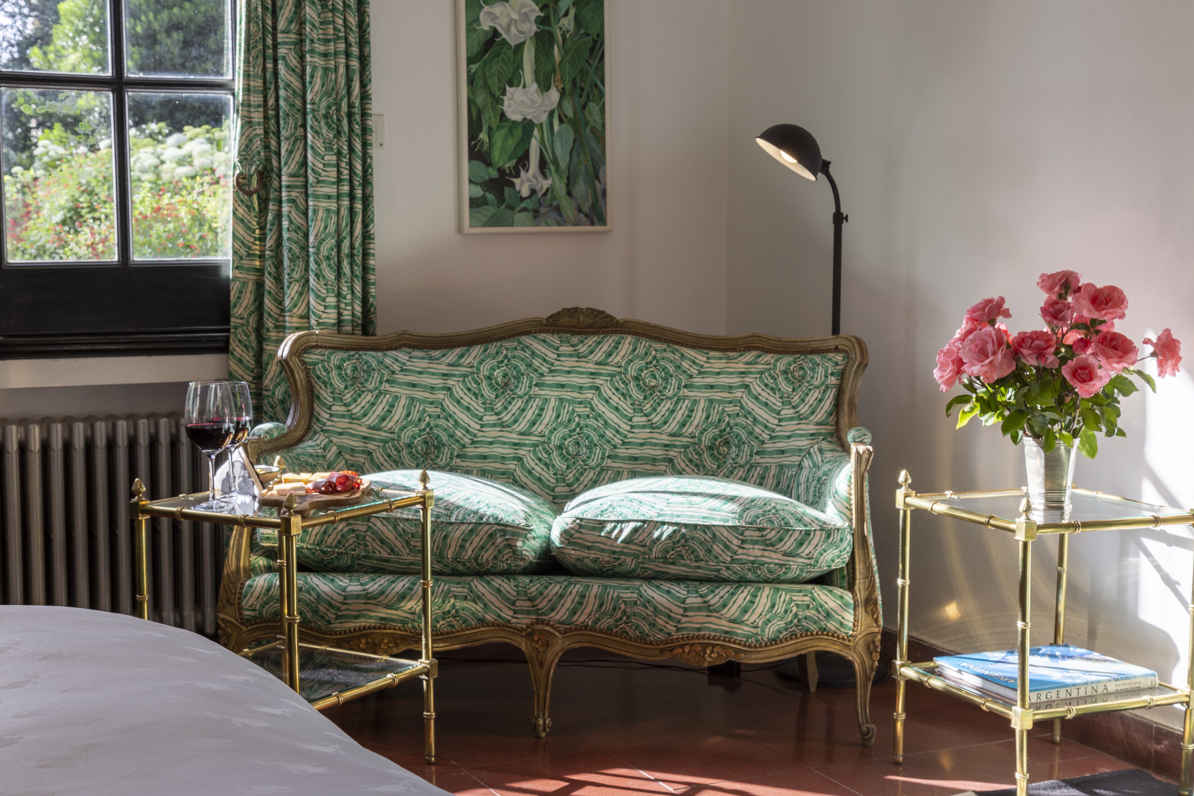 Um quarto com um sofá estampado, uma mesa pequena com um copo, um vaso com flores e um radiador.