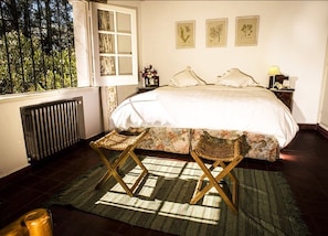 Une chambre avec un grand lit, une fenêtre donnant sur des arbres, un radiateur et une table de chevet avec une lampe.