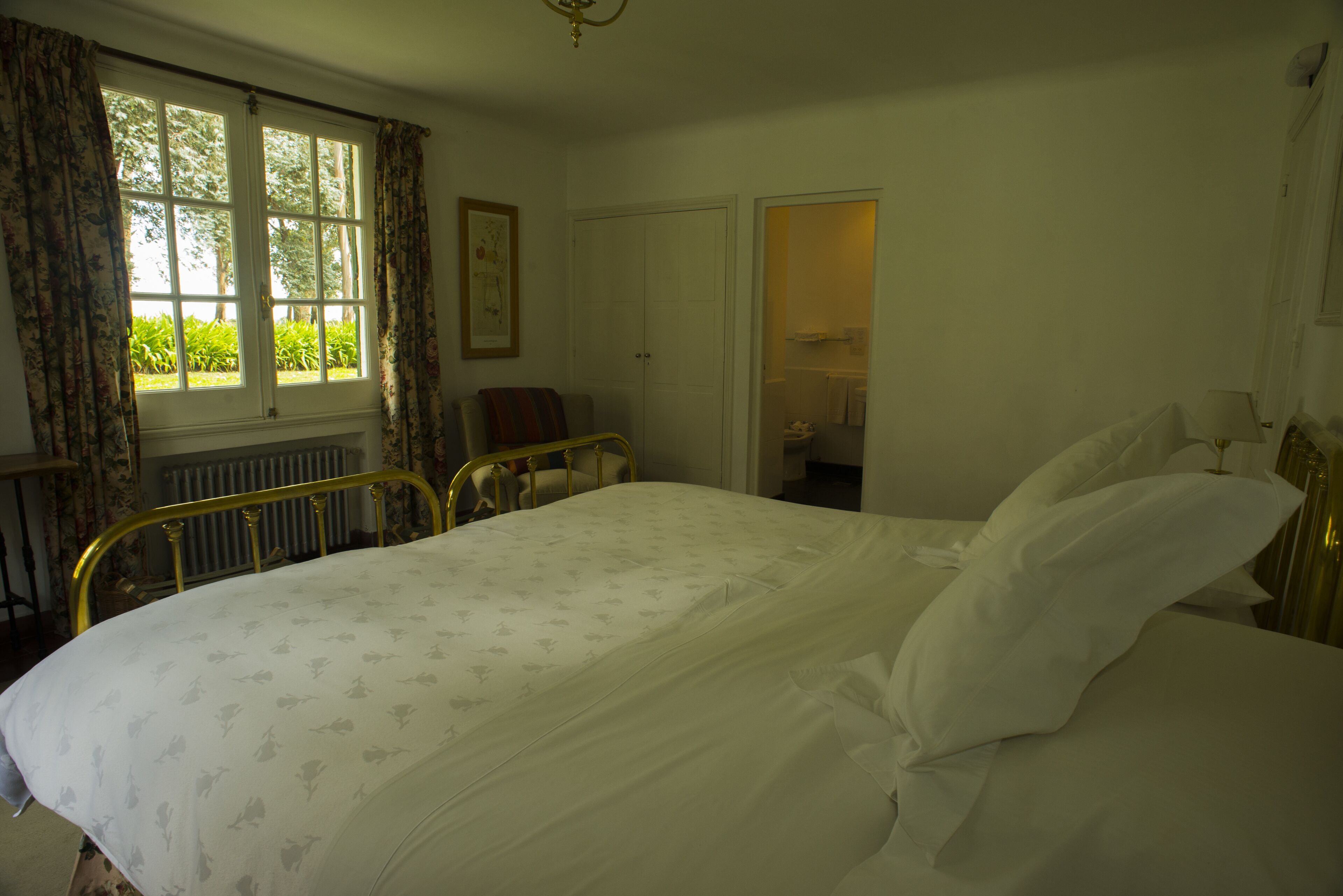 Une chambre à coucher comprenant un lit, des rideaux, un radiateur, une chaise et une porte donnant sur une salle de bain.