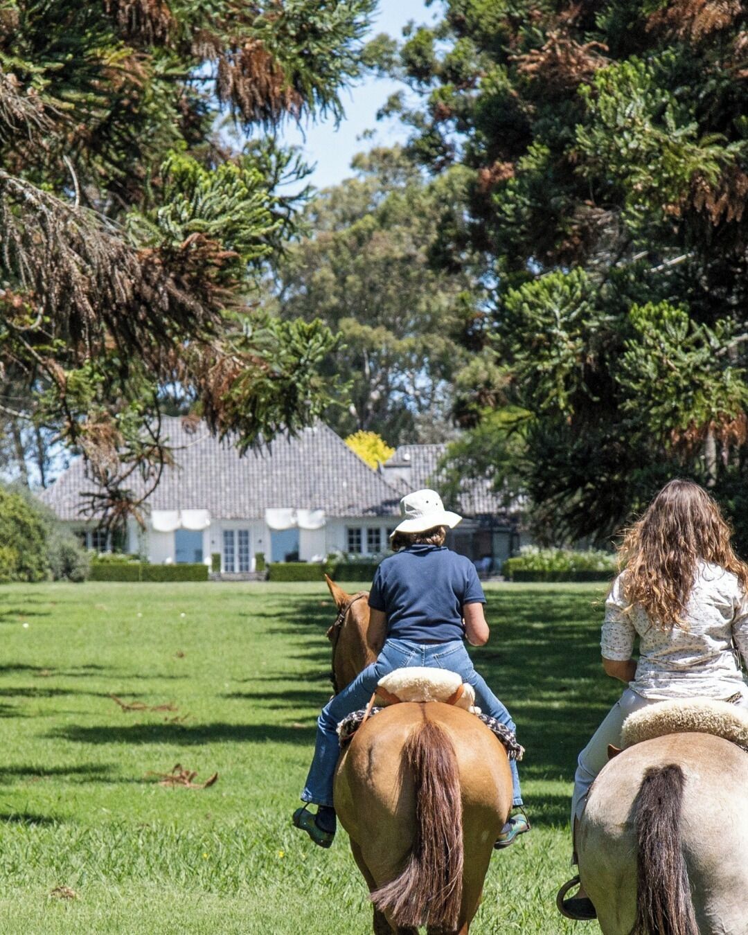 Two people riding horses in a grassy field with a house in the background.