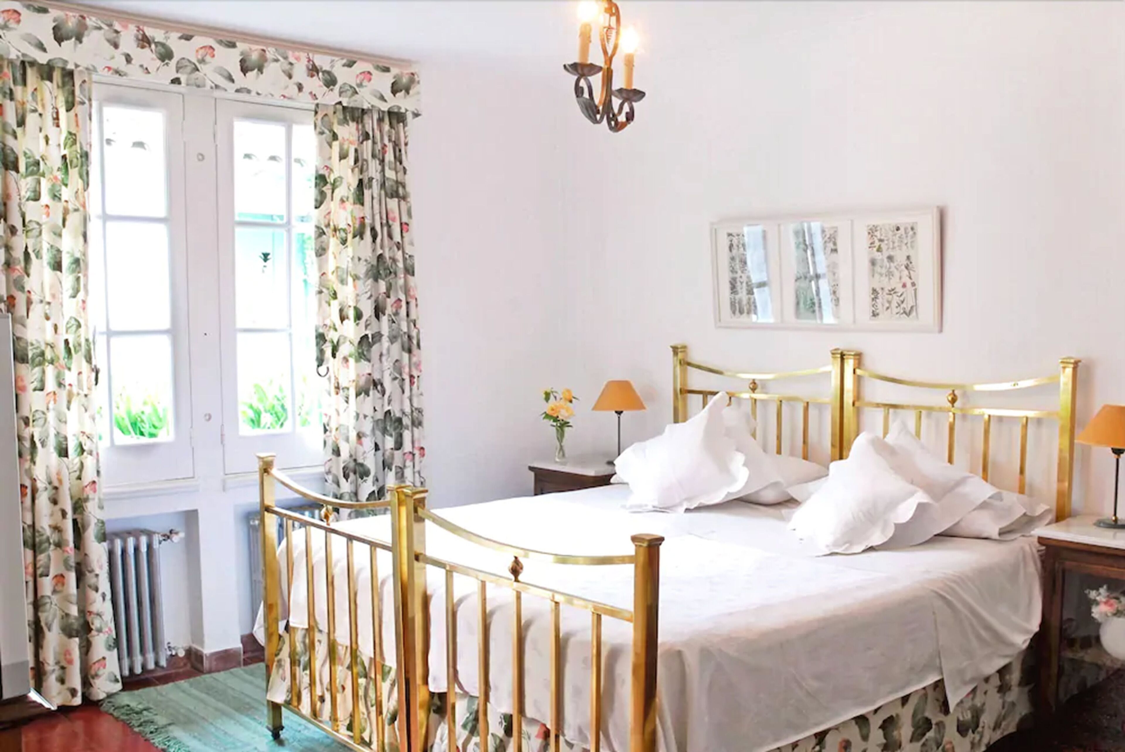 A bedroom with a bed, curtains, a chandelier, a nightstand, and a framed picture on the wall.