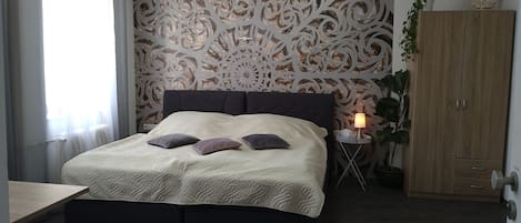 A bedroom with a large bed, a nightstand with a lamp, a wardrobe, and a decorative wall panel.