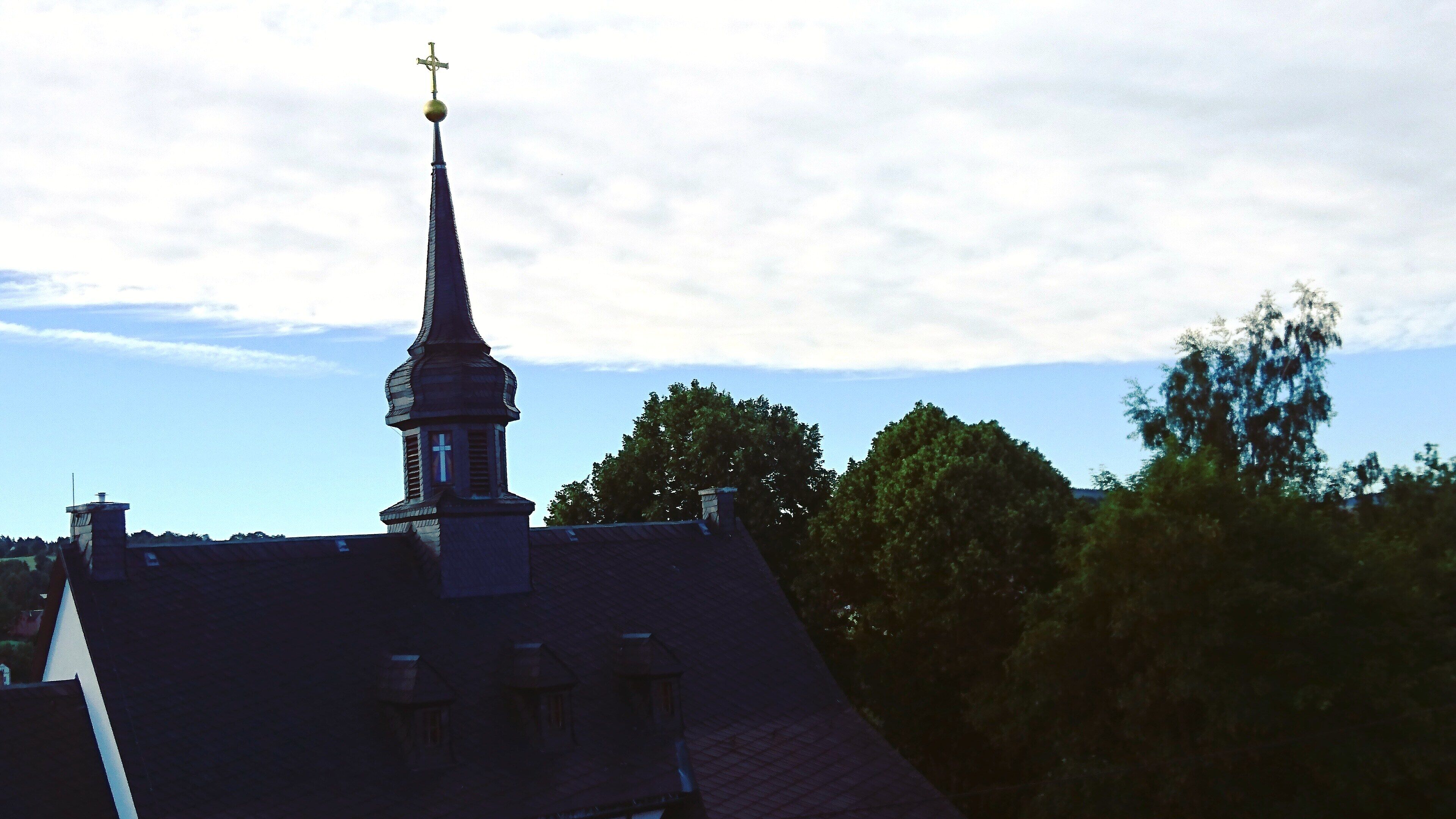 Een kerktoren met een kruis bovenop, omringd door bomen en een heldere lucht.