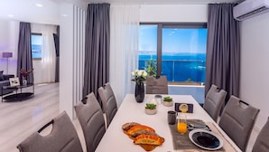 A modern dining area with a view of the sea, a white table set with bread, a vase with flowers, and a glass of orange juice.