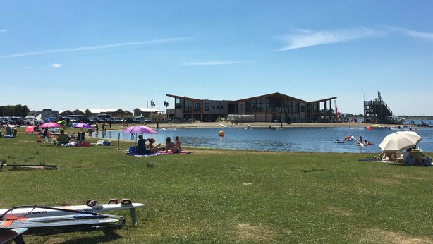 Ett område vid en sjö med en modern byggnad, gräsytor och människor som ägnar sig åt olika aktiviteter.