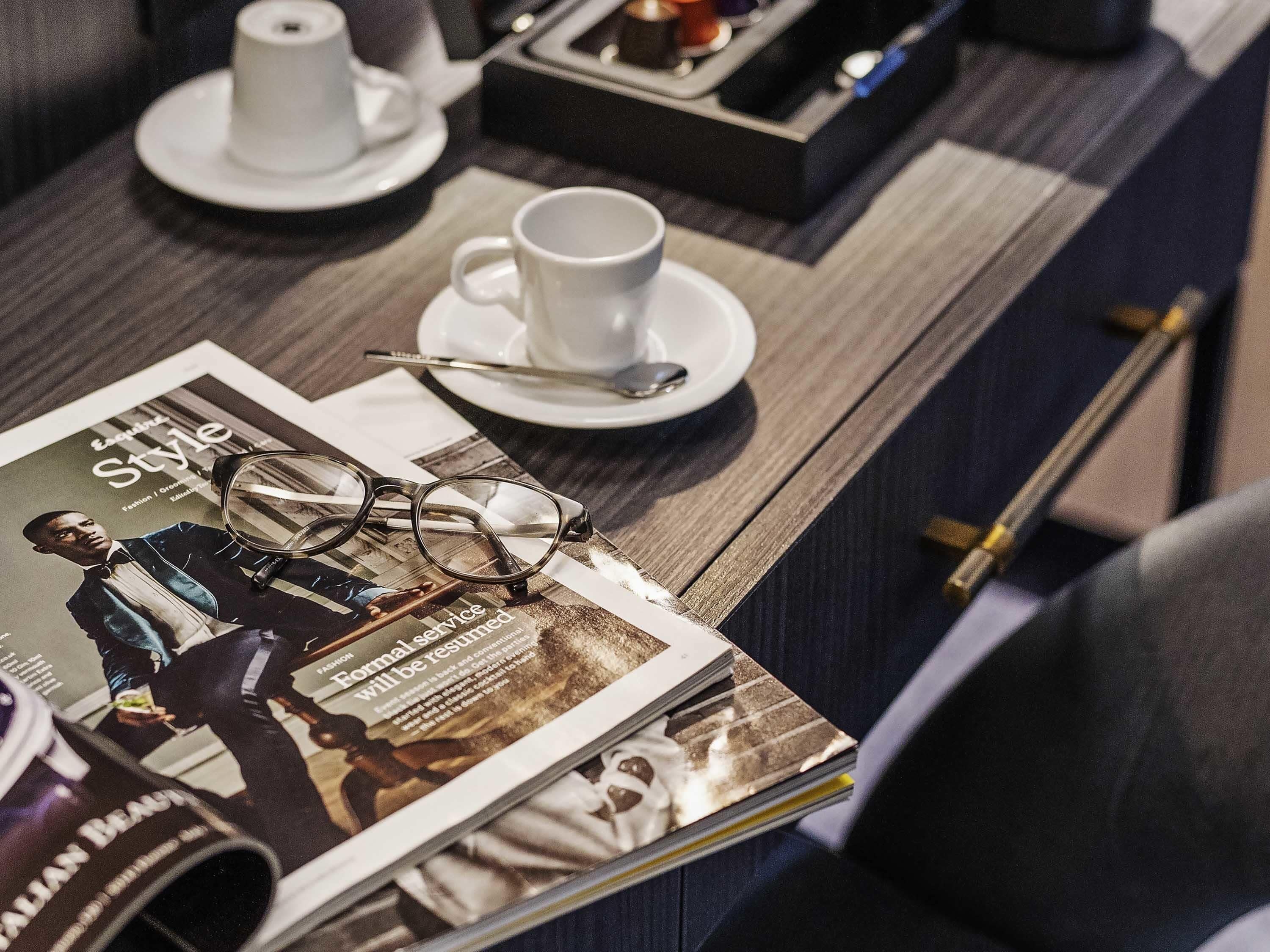 A magazine on a table, a pair of glasses, a cup, and a spoon.