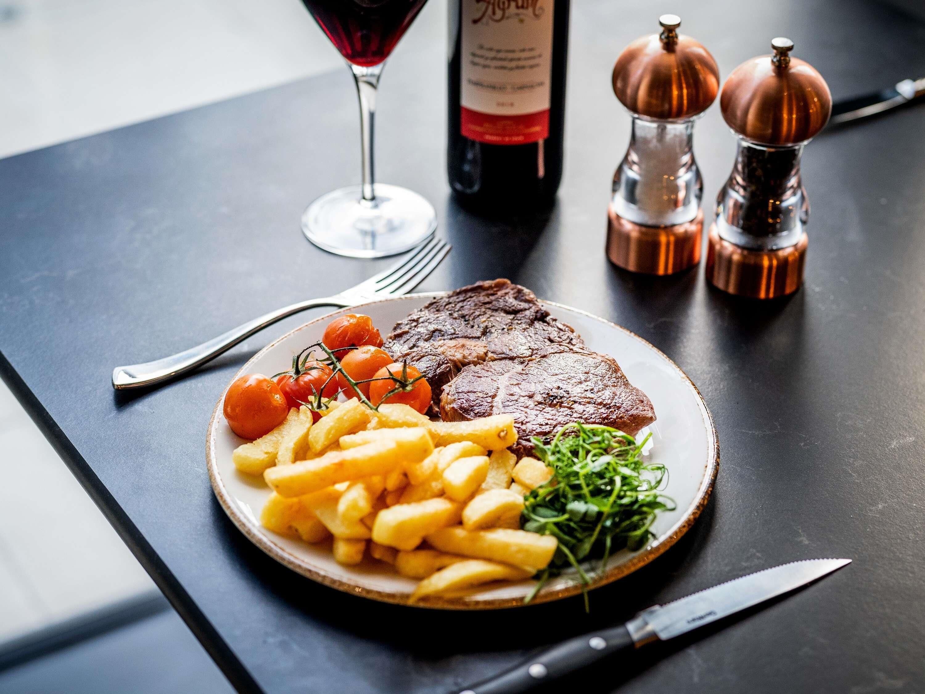 Un plato de comida con carne, papas fritas, verduras, una copa de vino y saleros y pimenteros sobre una mesa.