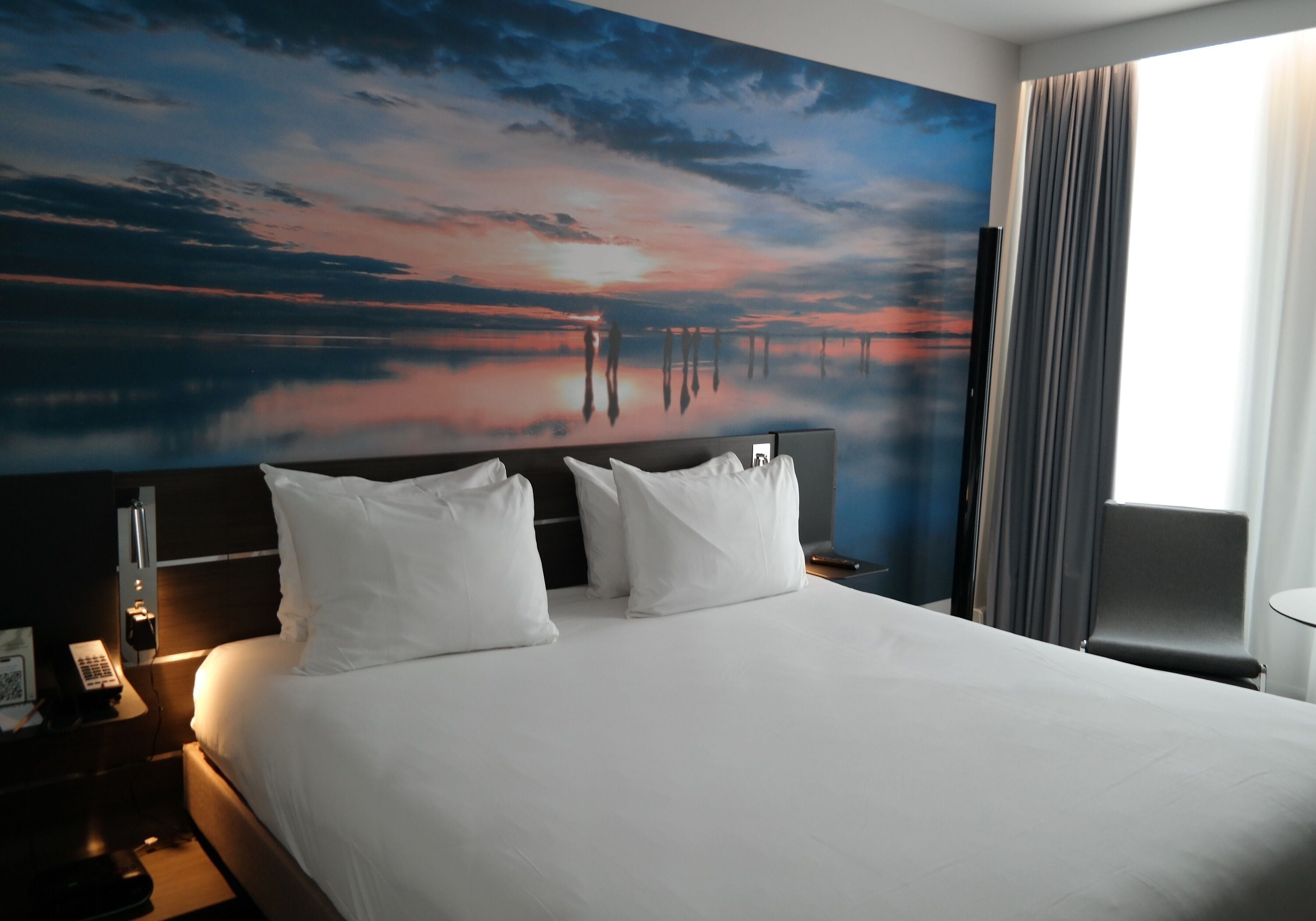 A hotel room with a large bed, a nightstand with a phone, and a painting of a sunset over water on the wall.