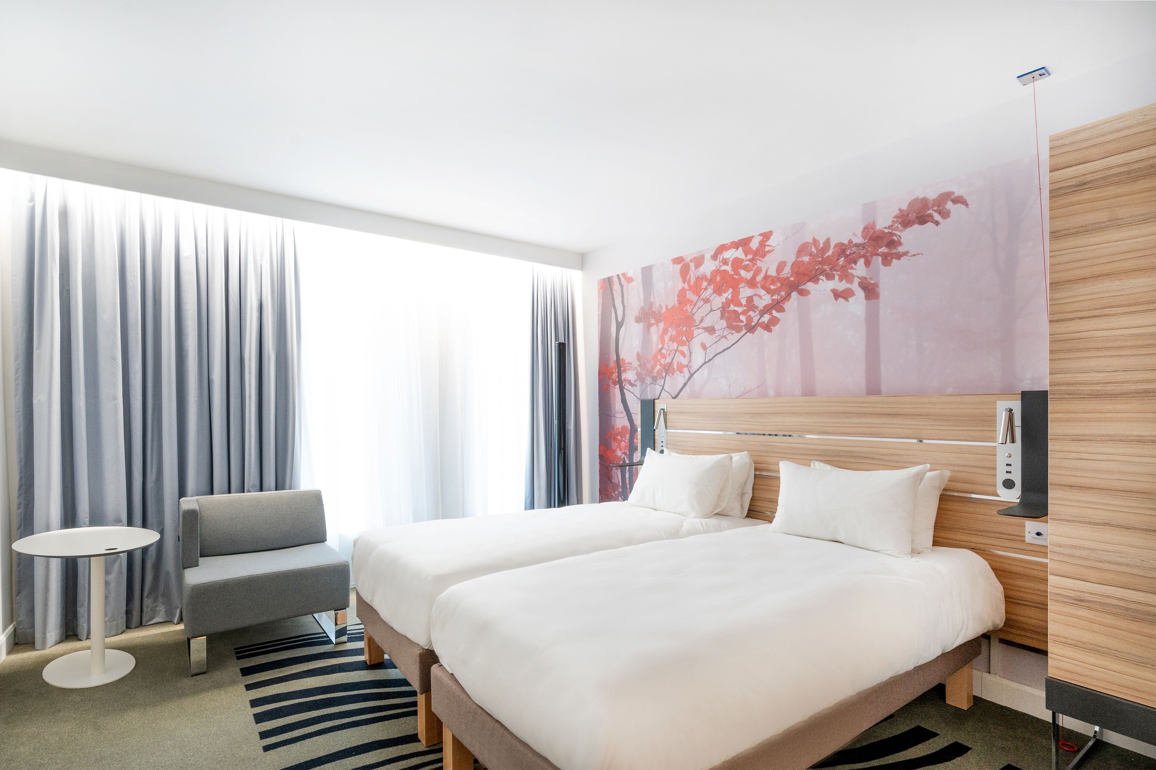 A hotel room with two beds, a grey sofa, a small round table, and a large window with curtains.