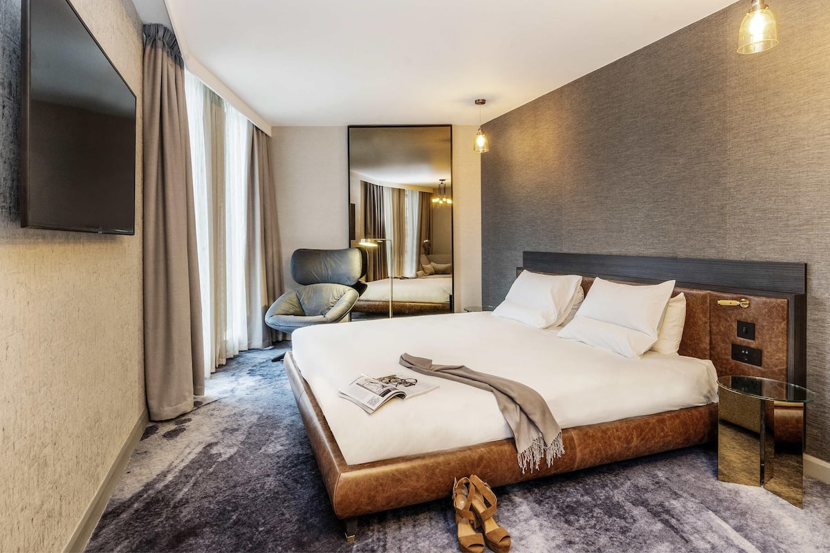 A modern hotel room with a large bed, a flat-screen TV, a chair, and a mirror.