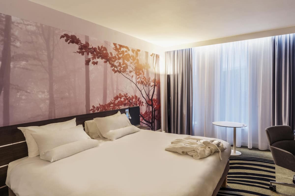 A hotel room with a large bed, a bedside table, a chair, and a view of the outdoors through sheer curtains.