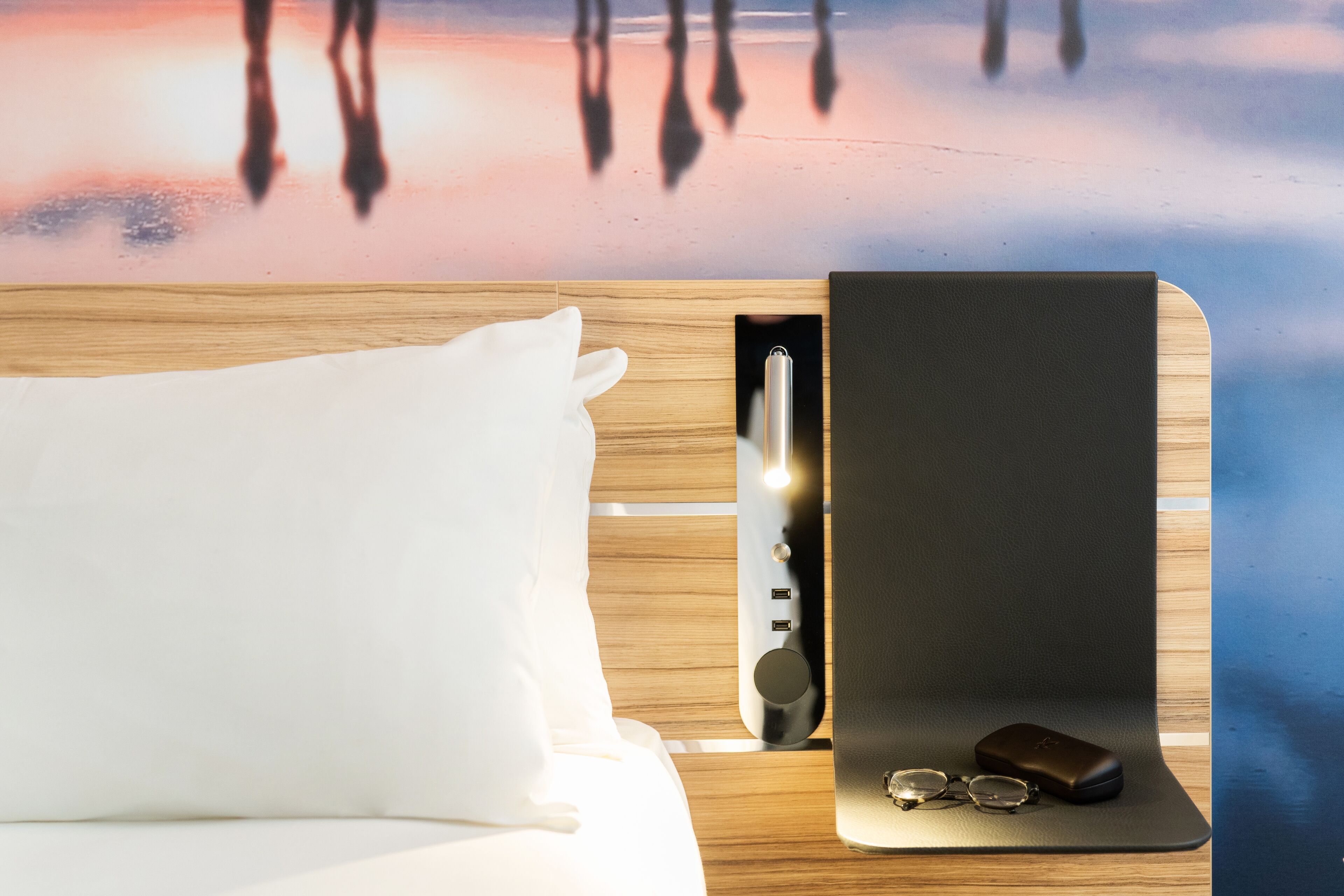 A neatly made bed with a white pillow, a bedside table with a black object and glasses, and a wooden headboard.