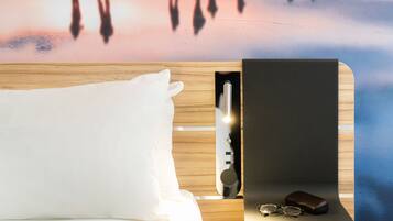 A neatly made bed with a white pillow, a bedside table with a black object and glasses, and a wooden headboard.