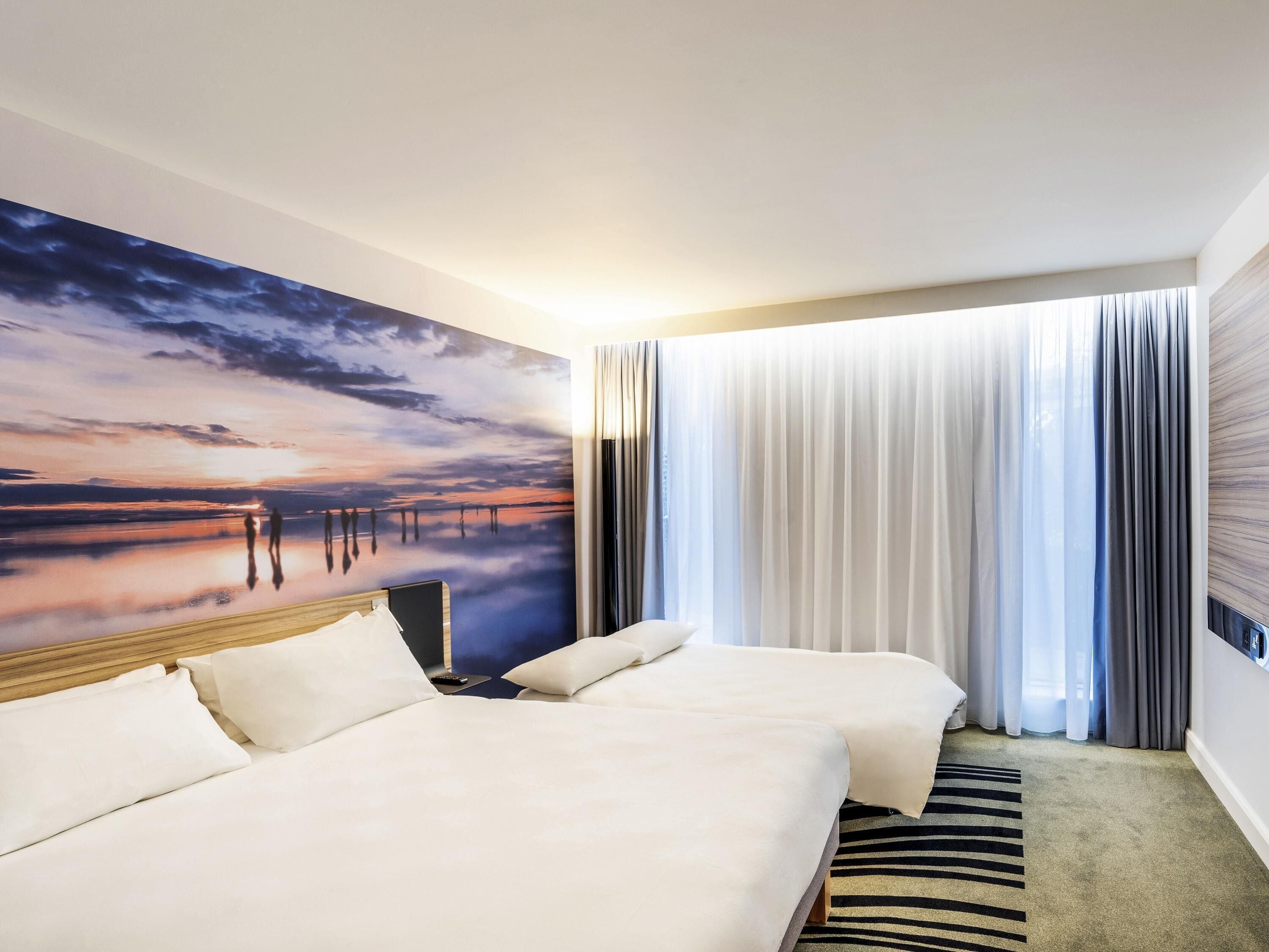 A hotel room with two beds, a large painting of a sunset, a TV, and a window with curtains.