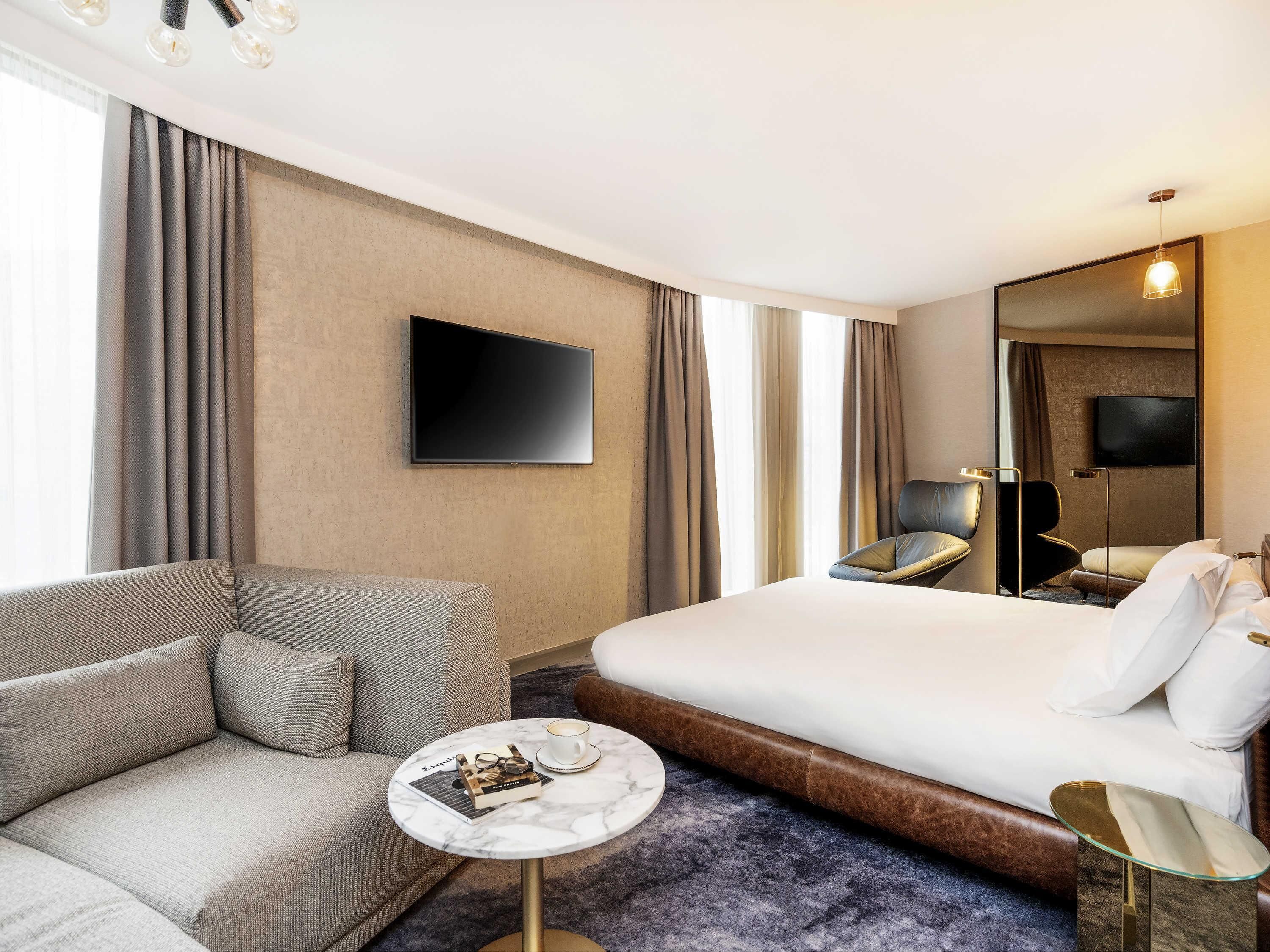 A hotel room with a sofa, a bed, a small round table with a book and a cup, a flat-screen TV, and a mirror.