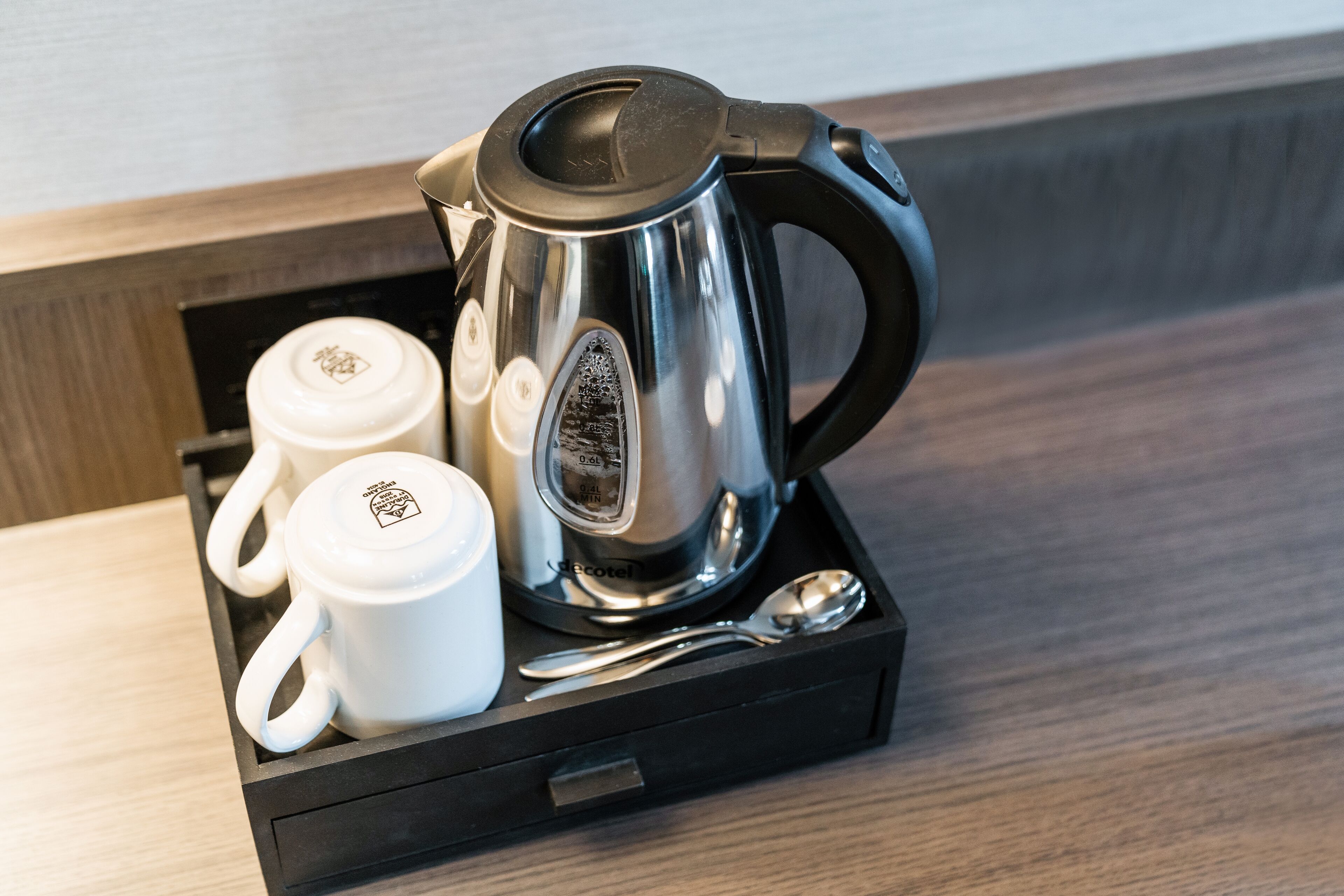 A stainless steel coffee maker, two white mugs, and a spoon on a black tray.