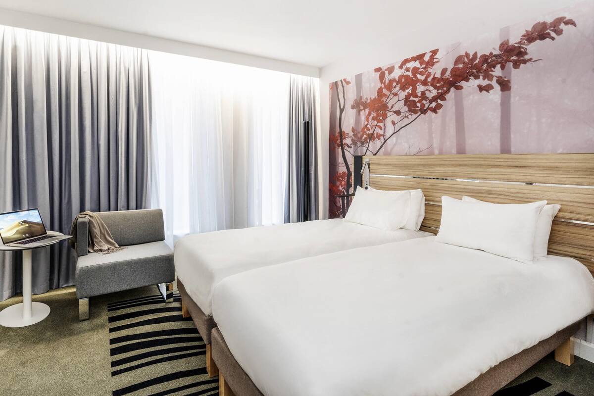 A hotel room with two beds, a desk with a laptop, a chair, and a large window with curtains.