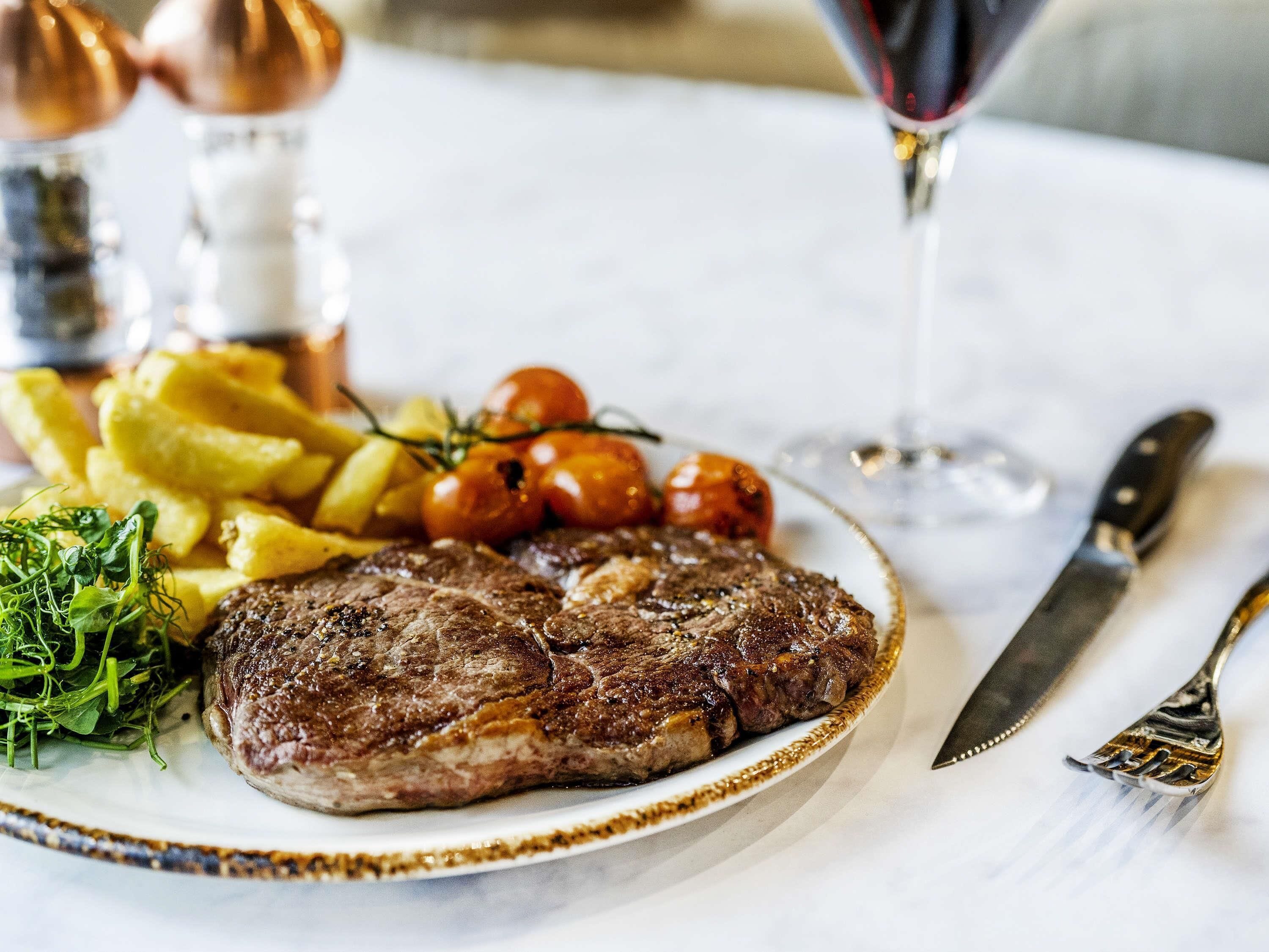 A plate of steak, fries, and cherry tomatoes on a table with a glass of red wine and cutlery.