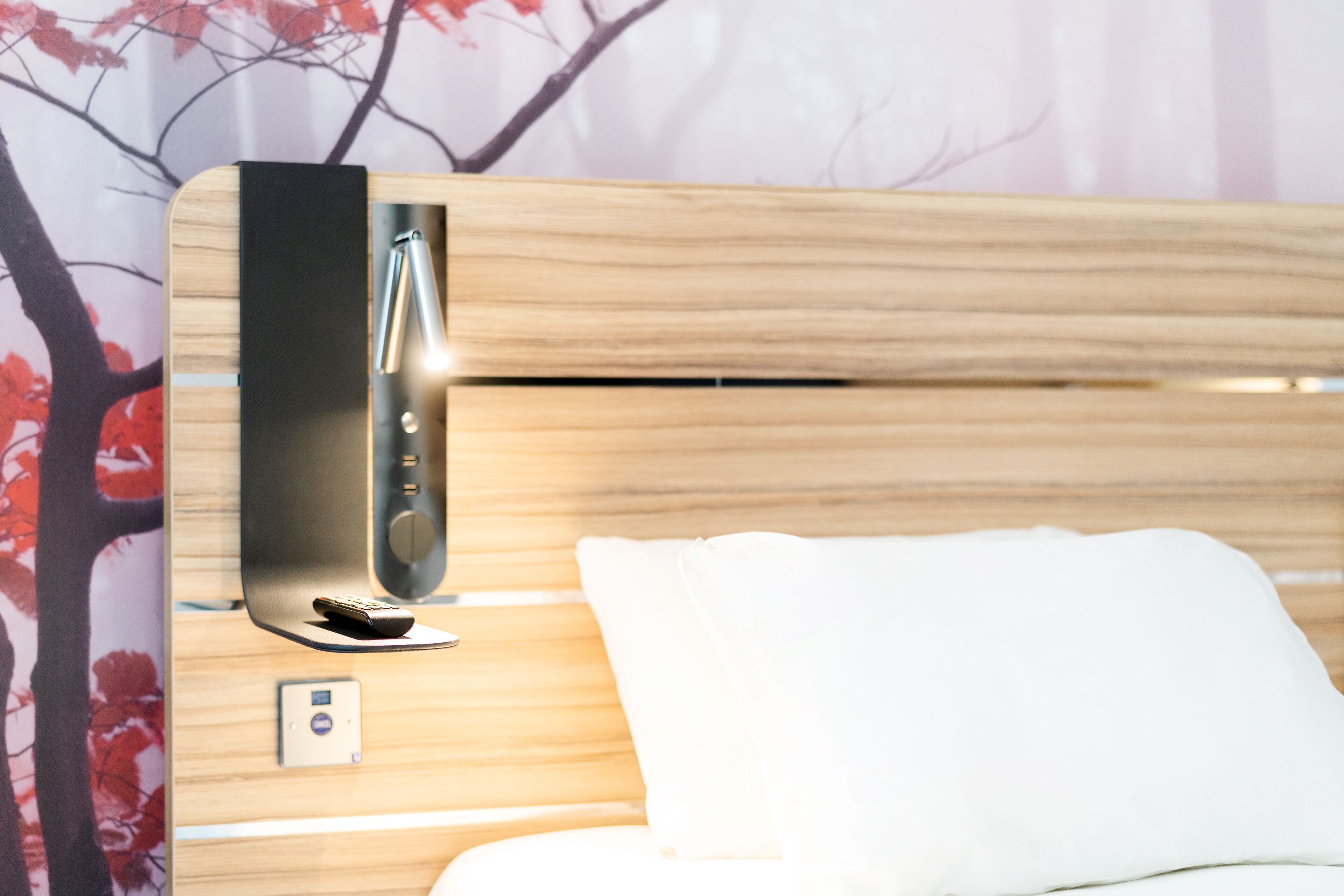 A modern hotel room with a wooden headboard, a bedside table with a remote control, and a wall-mounted thermostat.