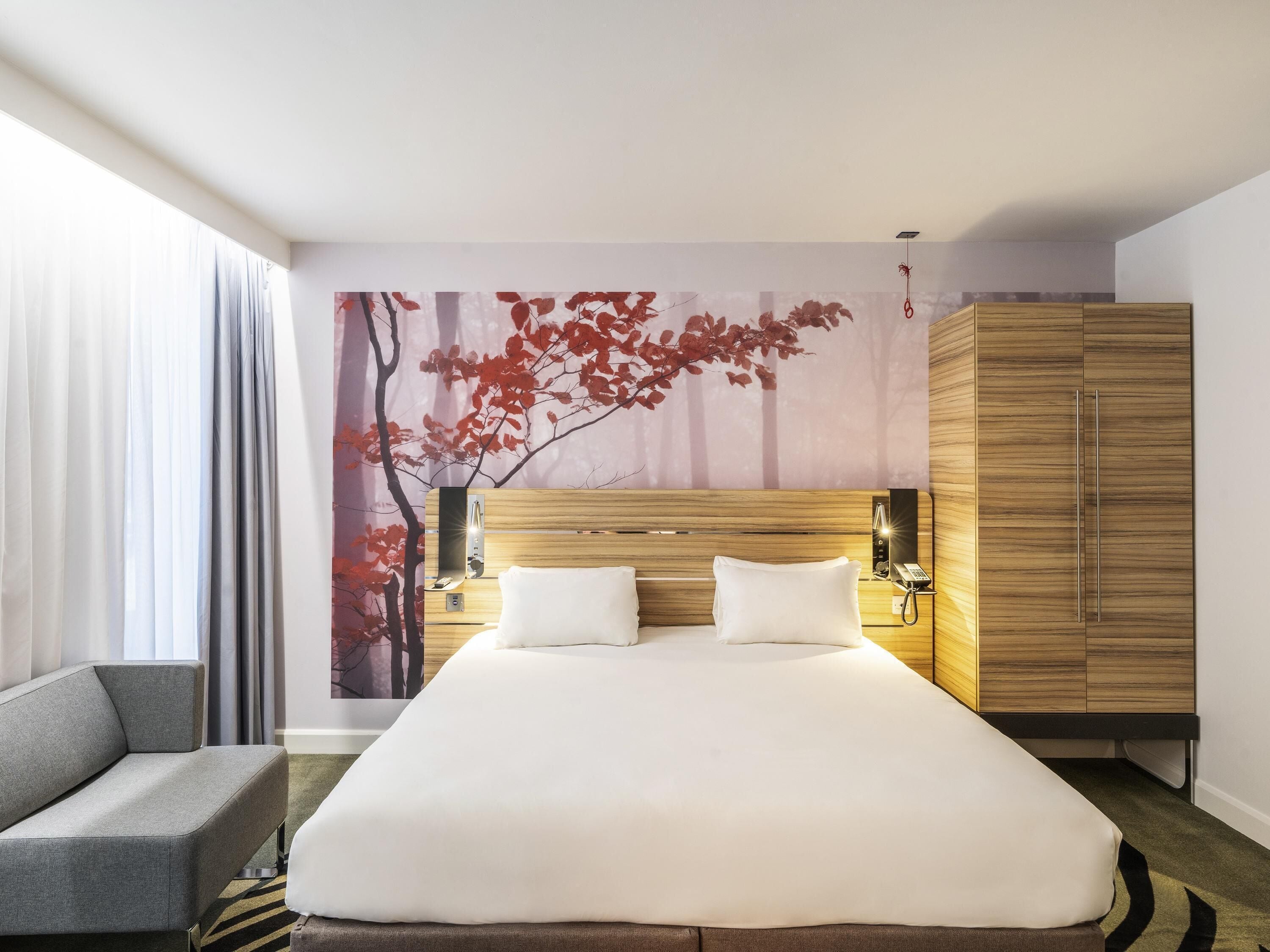 Habitación de hotel con una cama grande, un sofá gris, un armario y una pared decorada con un mural de temática natural.