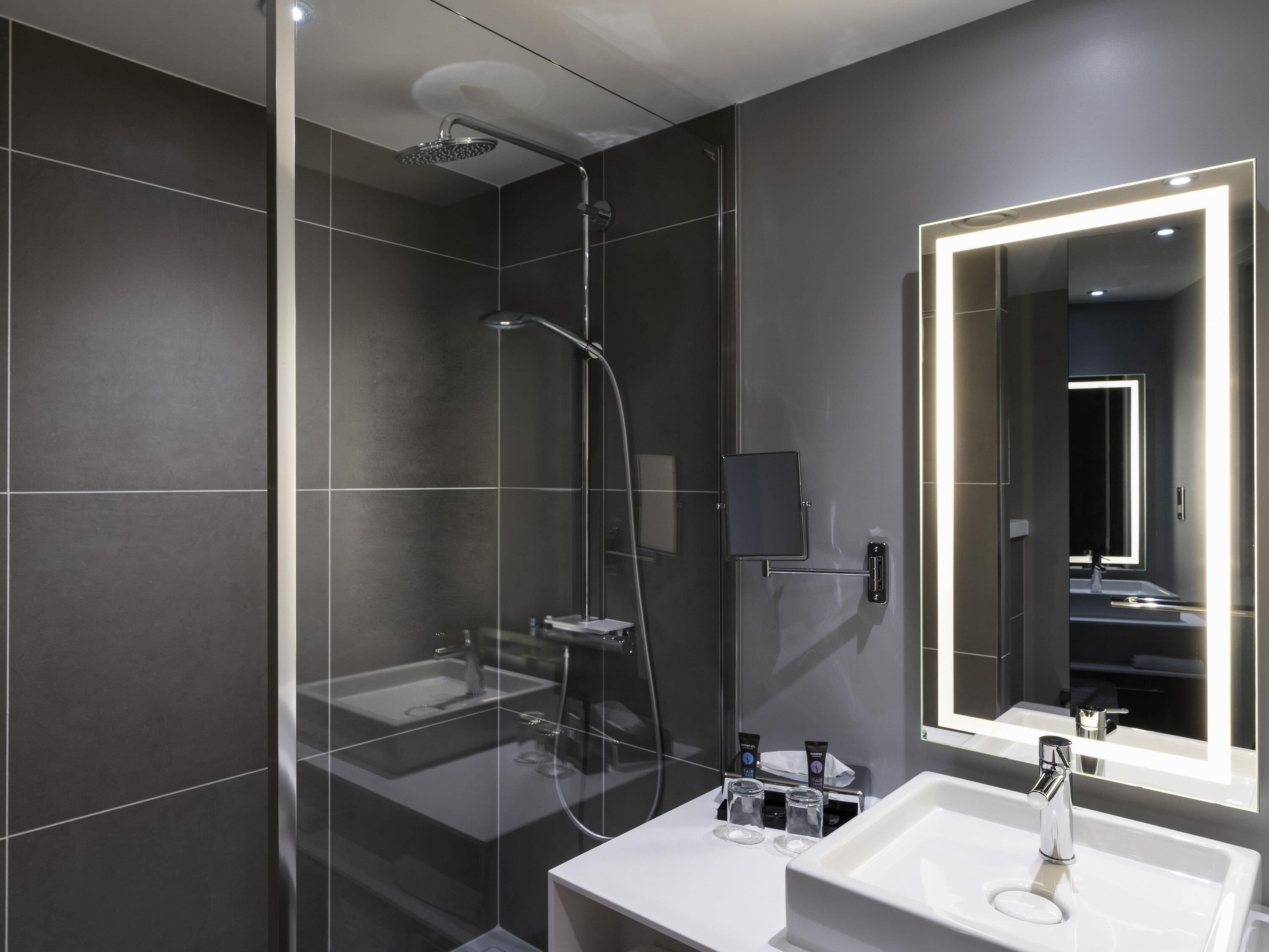 Une salle de bain moderne dotée d’une douche à parois de verre, d’un lavabo blanc et d’un grand miroir avec éclairage intégré.