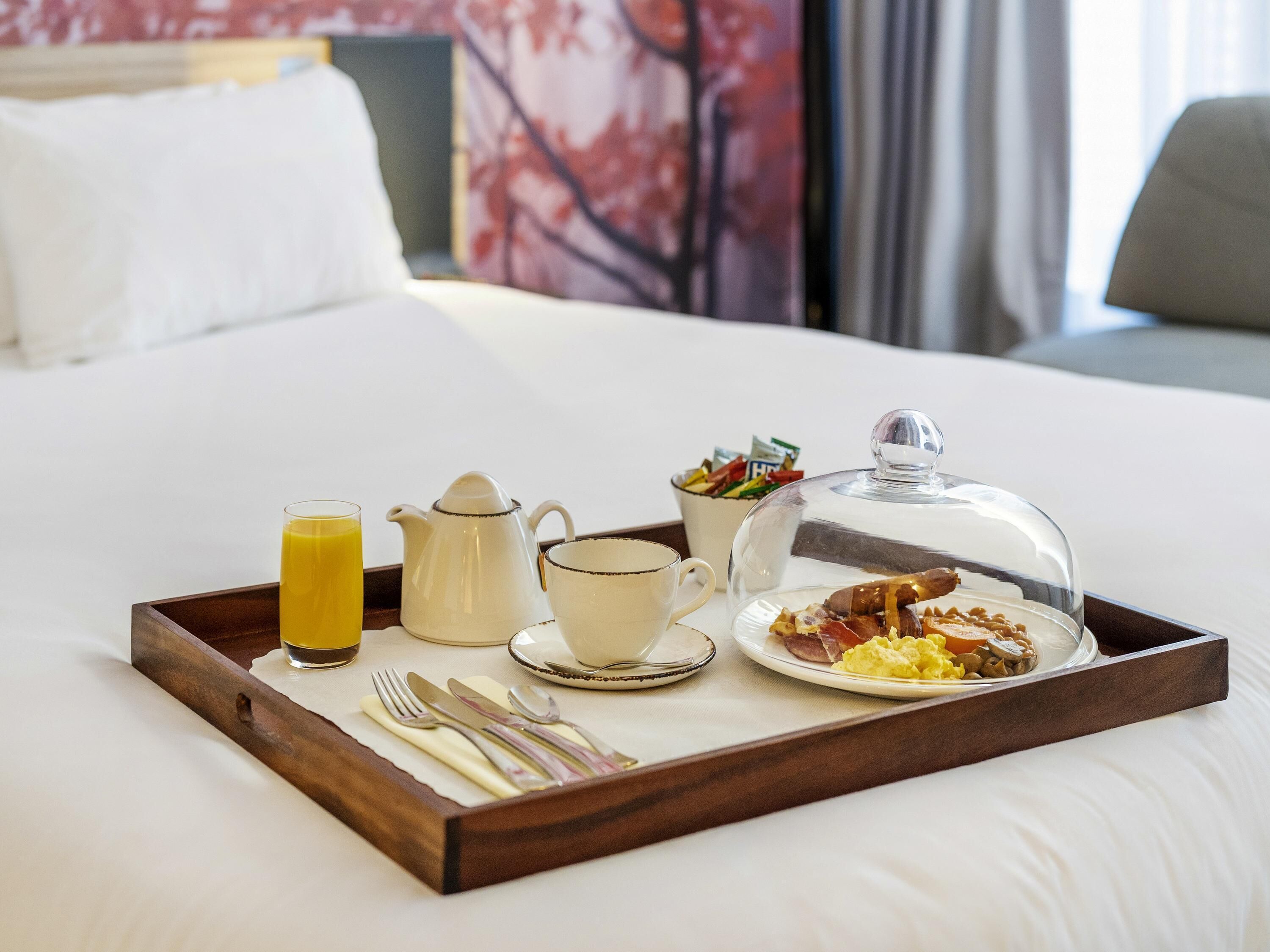 A breakfast tray on a bed with a glass of orange juice, a teapot, a cup, a bowl of cereal, and a plate with eggs and bacon.