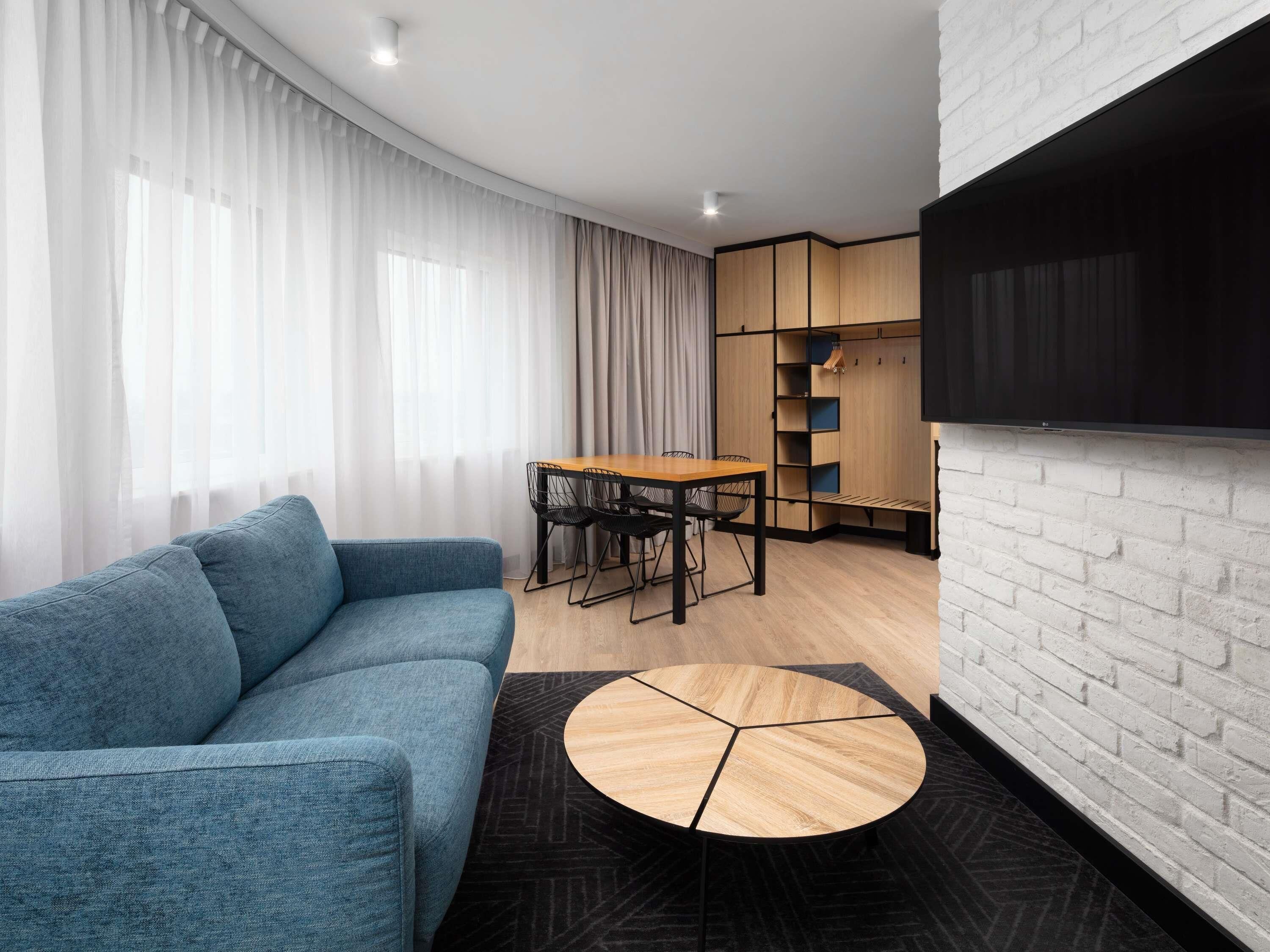 A modern living room with a blue sofa, a wooden dining table with chairs, a round coffee table, and a TV mounted on a white brick wall.